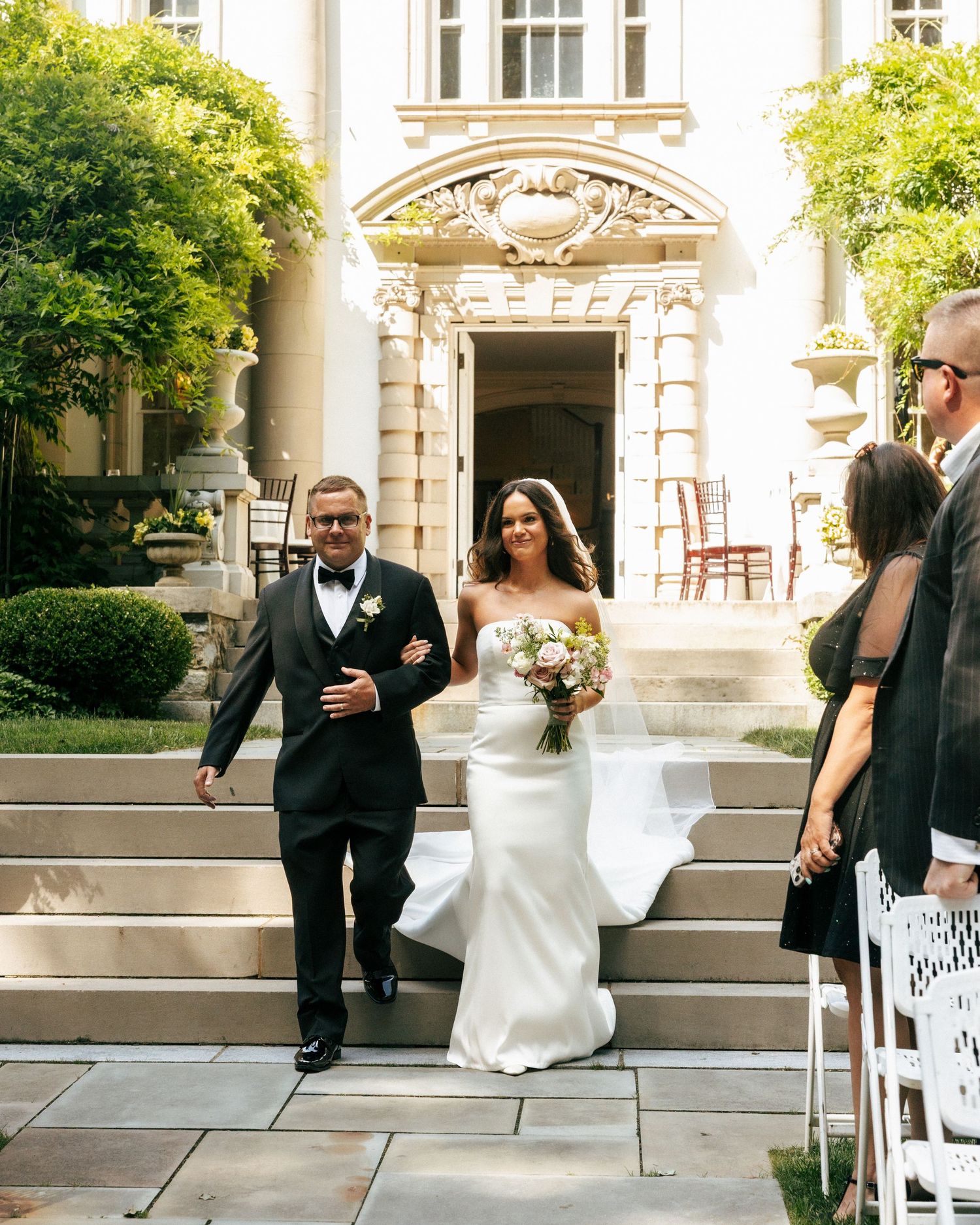 Bride in white gown walks down outdoor steps with escort at elegant mansion wedding ceremony.