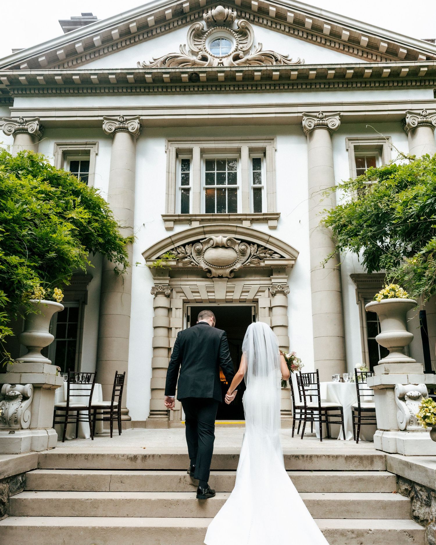 Bride and groom walk hand-in-hand toward an elegant neoclassical mansion entrance on their wedding day.