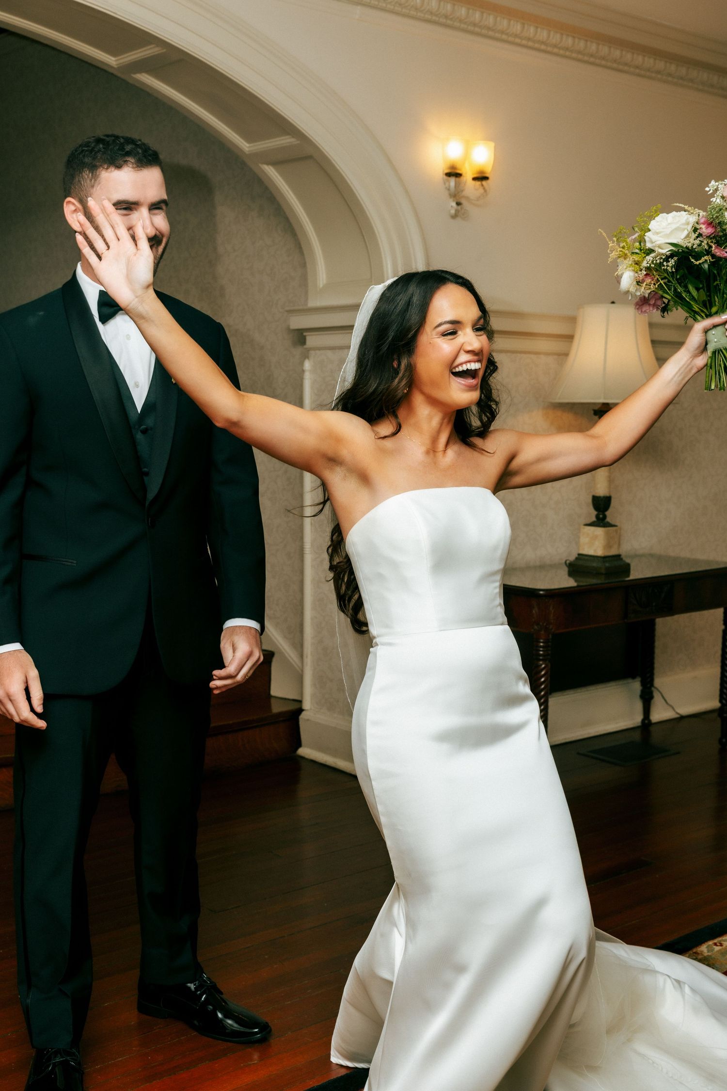 Joyful bride in strapless gown raises bouquet while groom covers his smile during wedding celebration indoors.