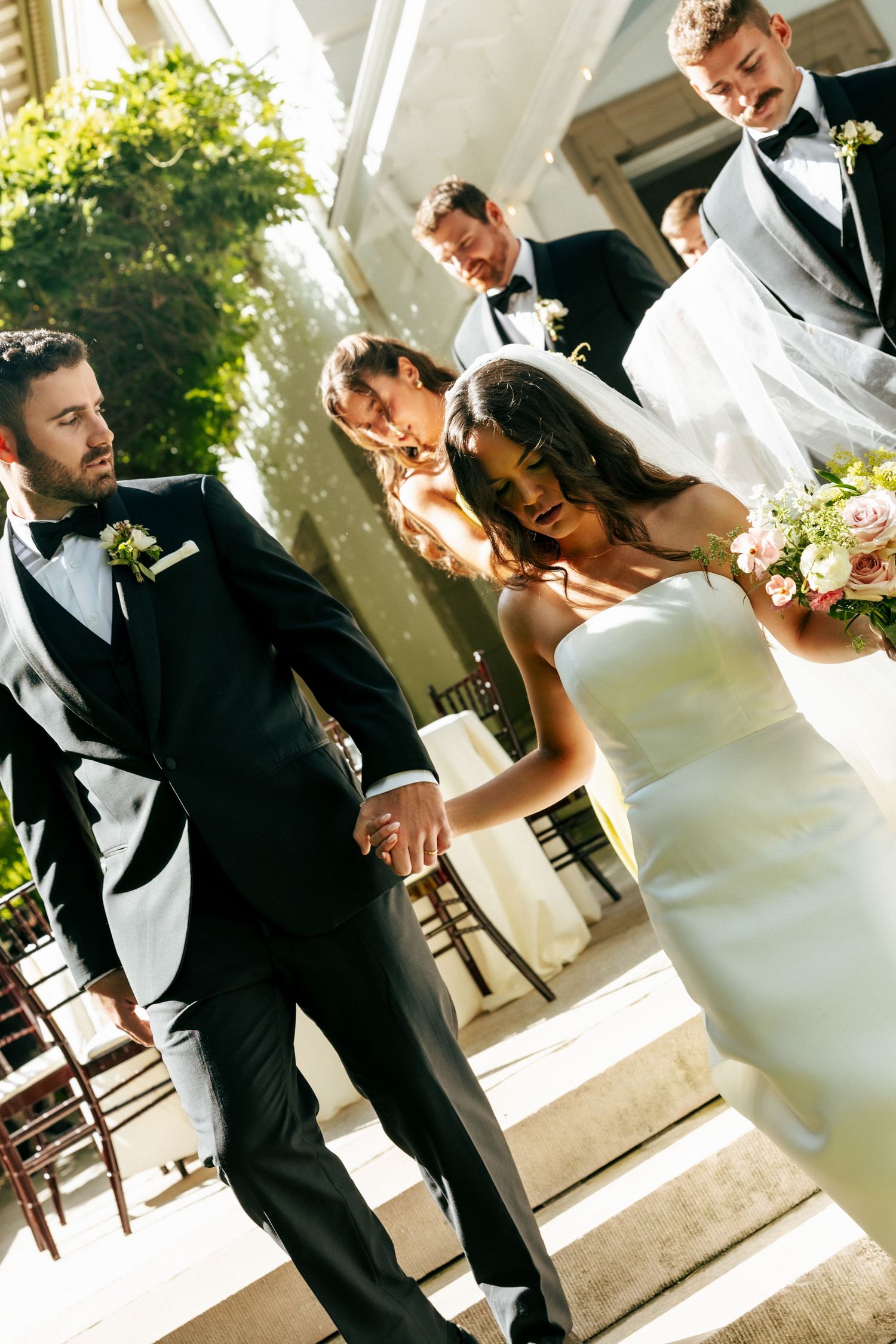 Bride and groom hold hands descending steps at outdoor wedding ceremony with groomsmen in black tuxedos behind them.