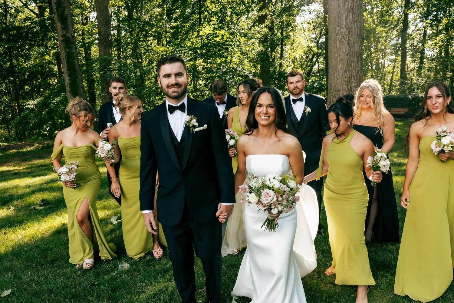 Wedding party walks outdoors, bride in white gown, groom in black tux, bridesmaids in yellow-green dresses, groomsmen in black suits.
