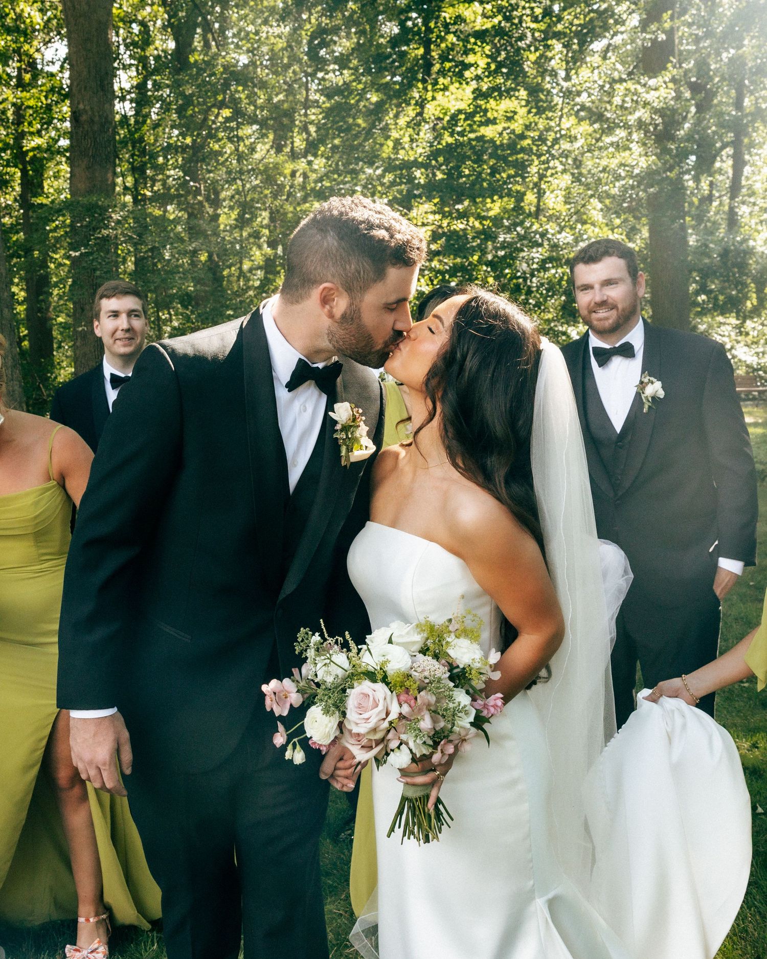 A bride and groom share a kiss outdoors, surrounded by their wedding party in a lush green forest setting.