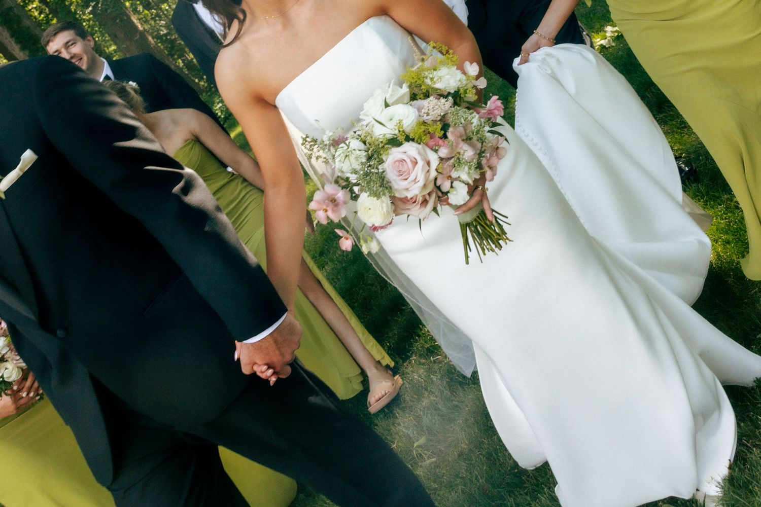 Bride in white gown holding pink and white bouquet standing with wedding party in formal attire outdoors.