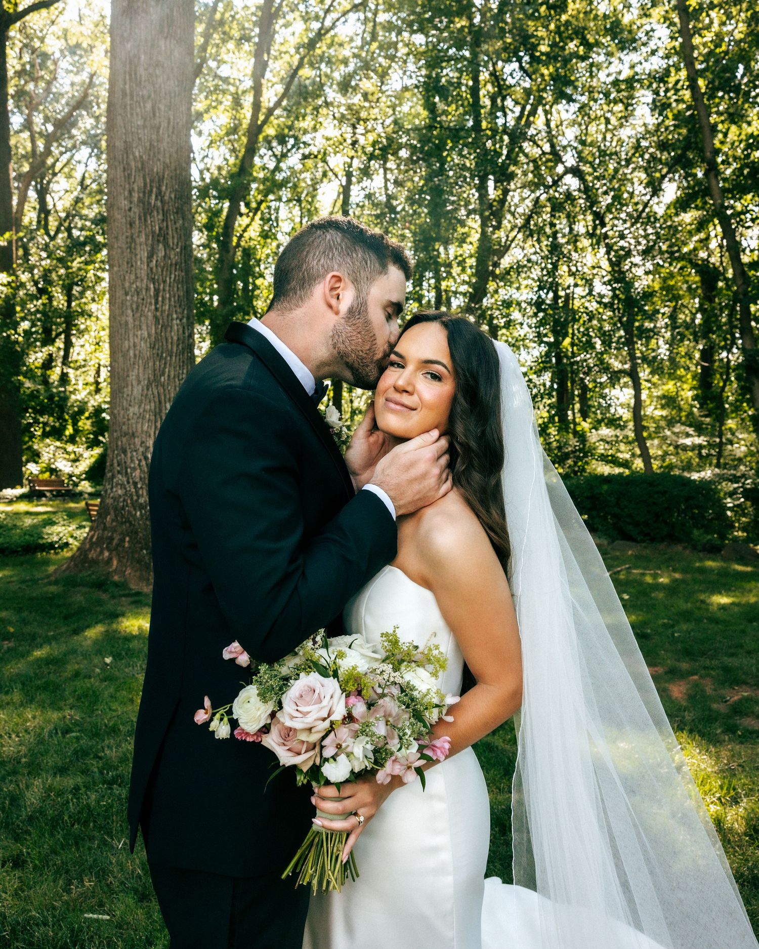 Bride in white gown with veil holds pink bouquet as groom in black tux kisses her cheek outdoors.
