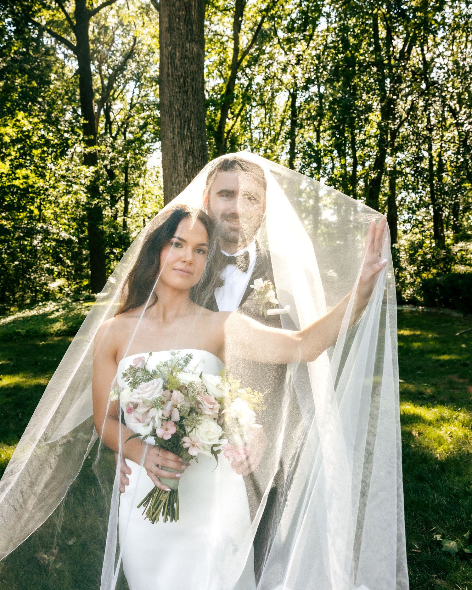 Bride and groom pose under flowing white veil outdoors, bride holding floral bouquet, surrounded by lush green trees.