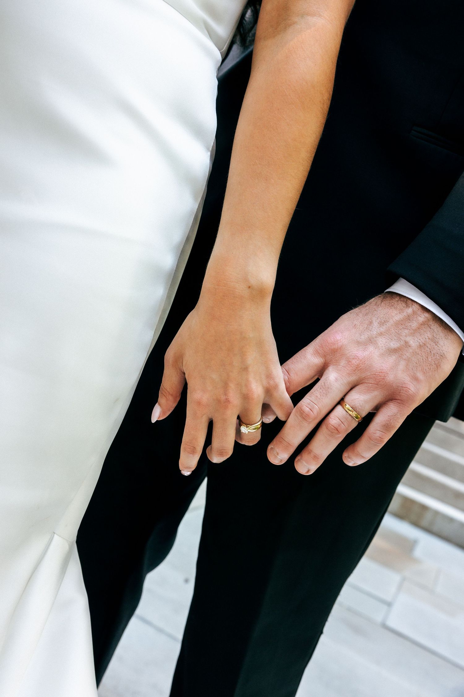 Newlywed couple holding hands showing matching gold wedding bands, bride in white gown and groom in black suit.