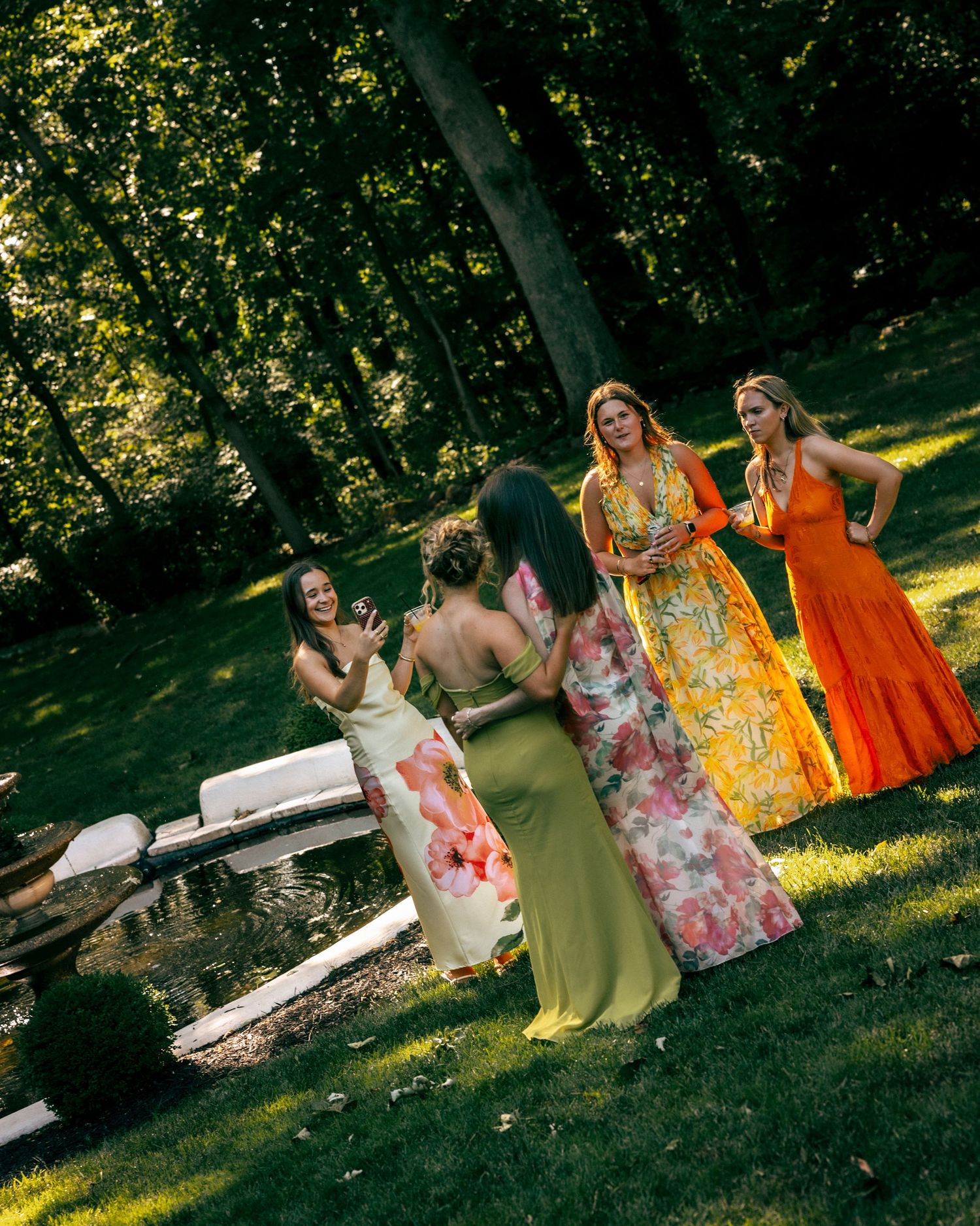Five women in colorful floral and solid maxi dresses socializing outdoors near a pond in a lush green garden.