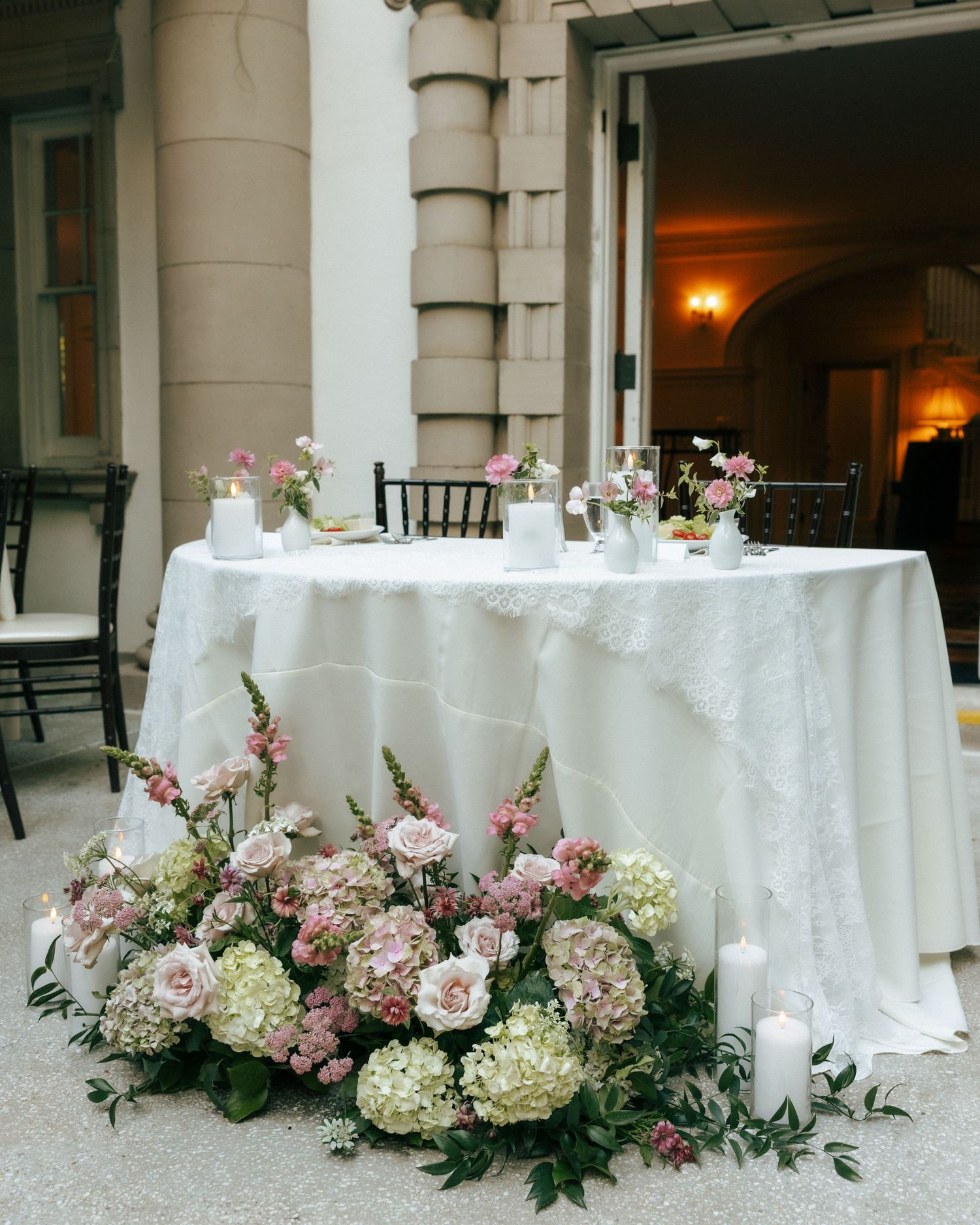 Elegant wedding sweetheart table with white lace linen, surrounded by lush floral arrangement of pink roses and white hydrangeas.