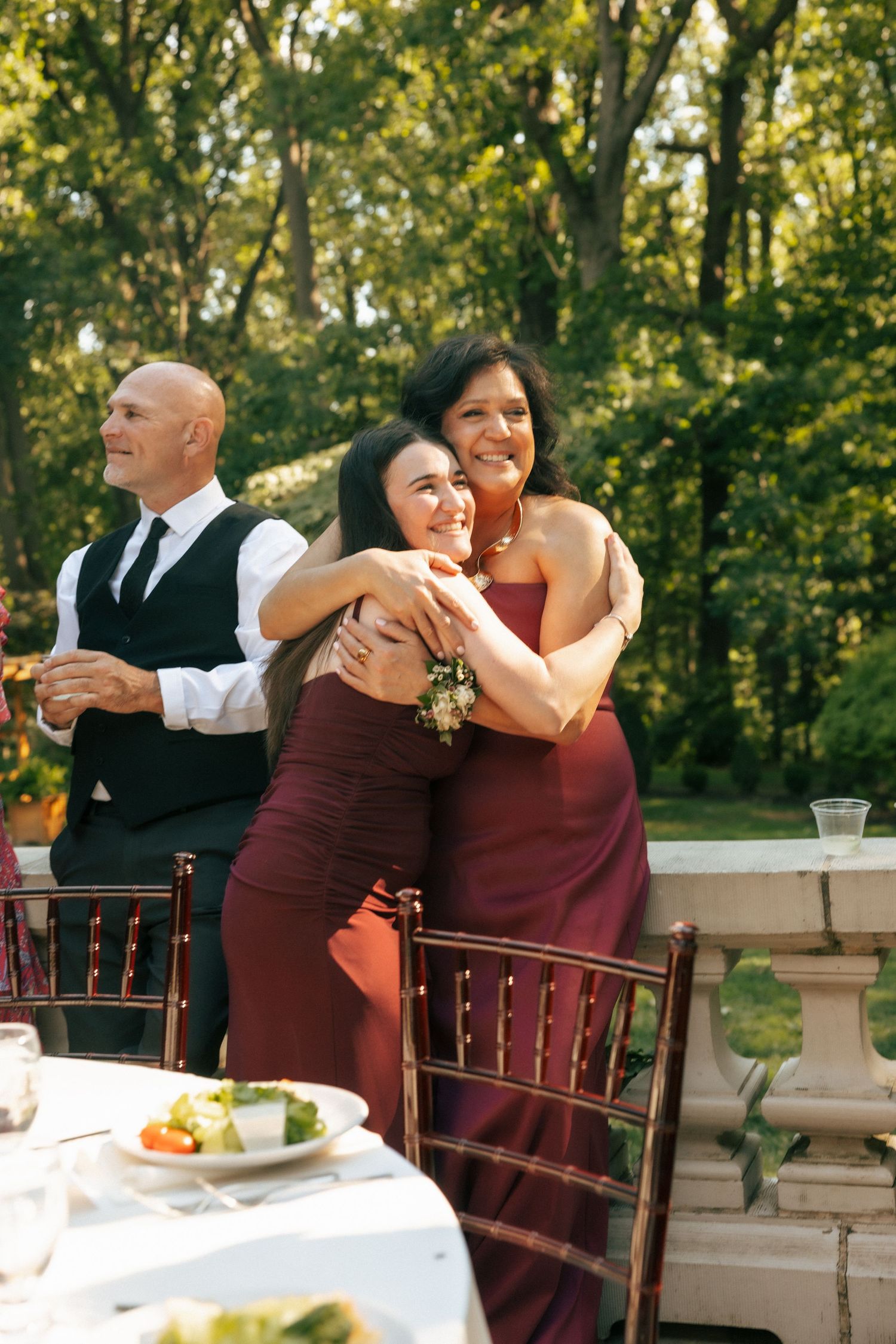 Two women in burgundy bridesmaid dresses share a warm hug at an outdoor wedding reception.