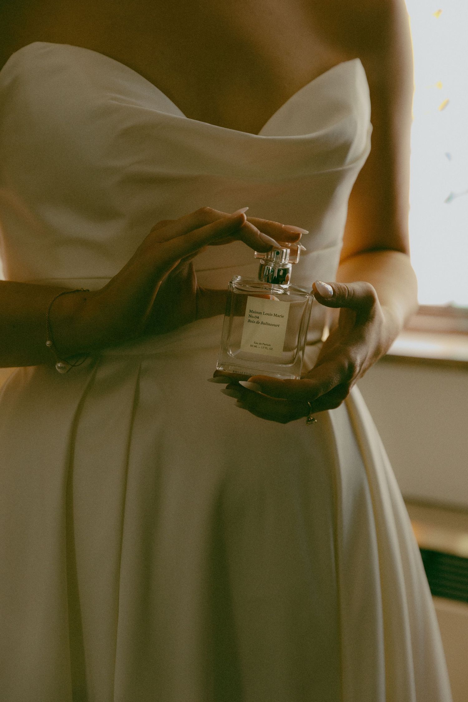 Close-up of hands holding a perfume bottle against a white wedding dress.