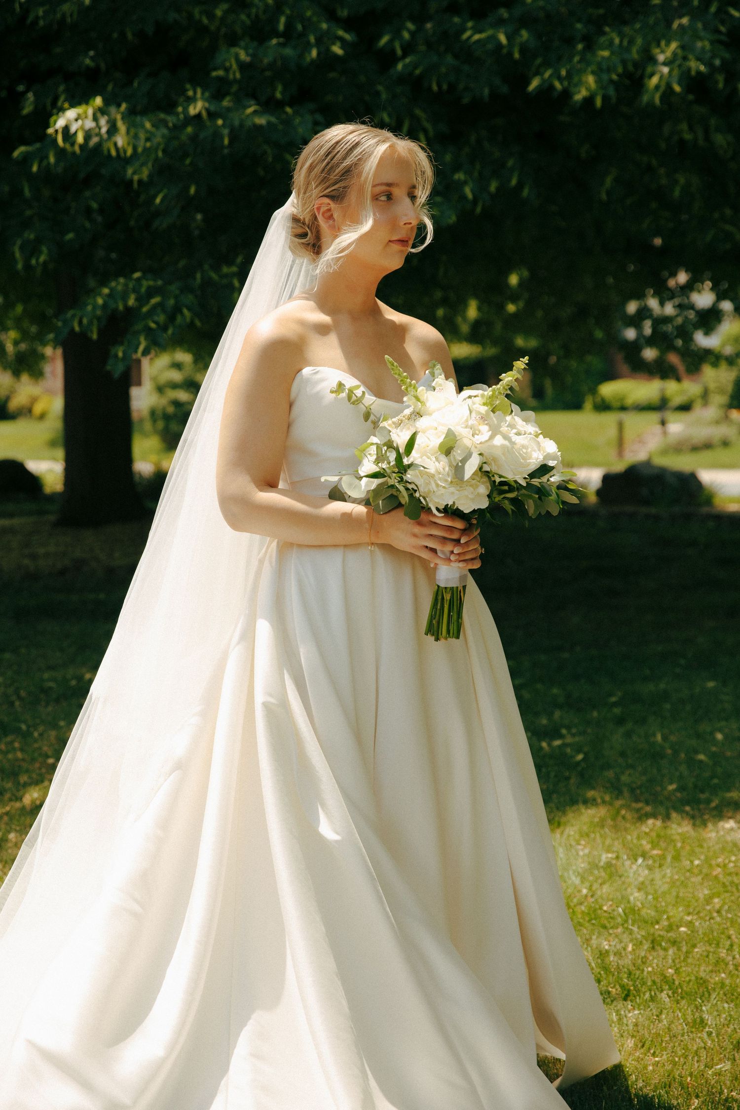 A bride in a classic white gown holds a bouquet of white flowers in a garden setting.