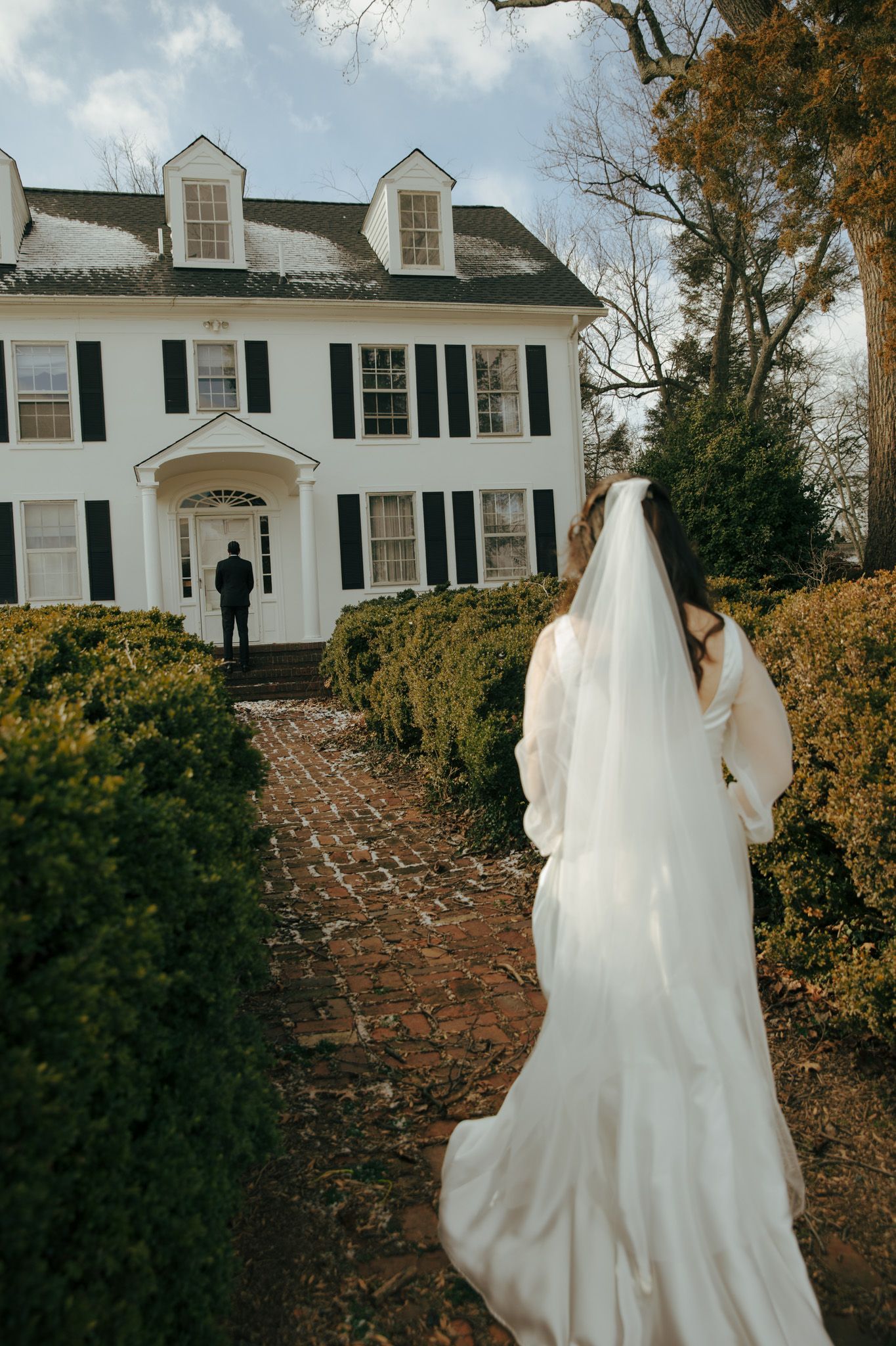 A bride approaches a classic white colonial house with black shutters on a brick path.