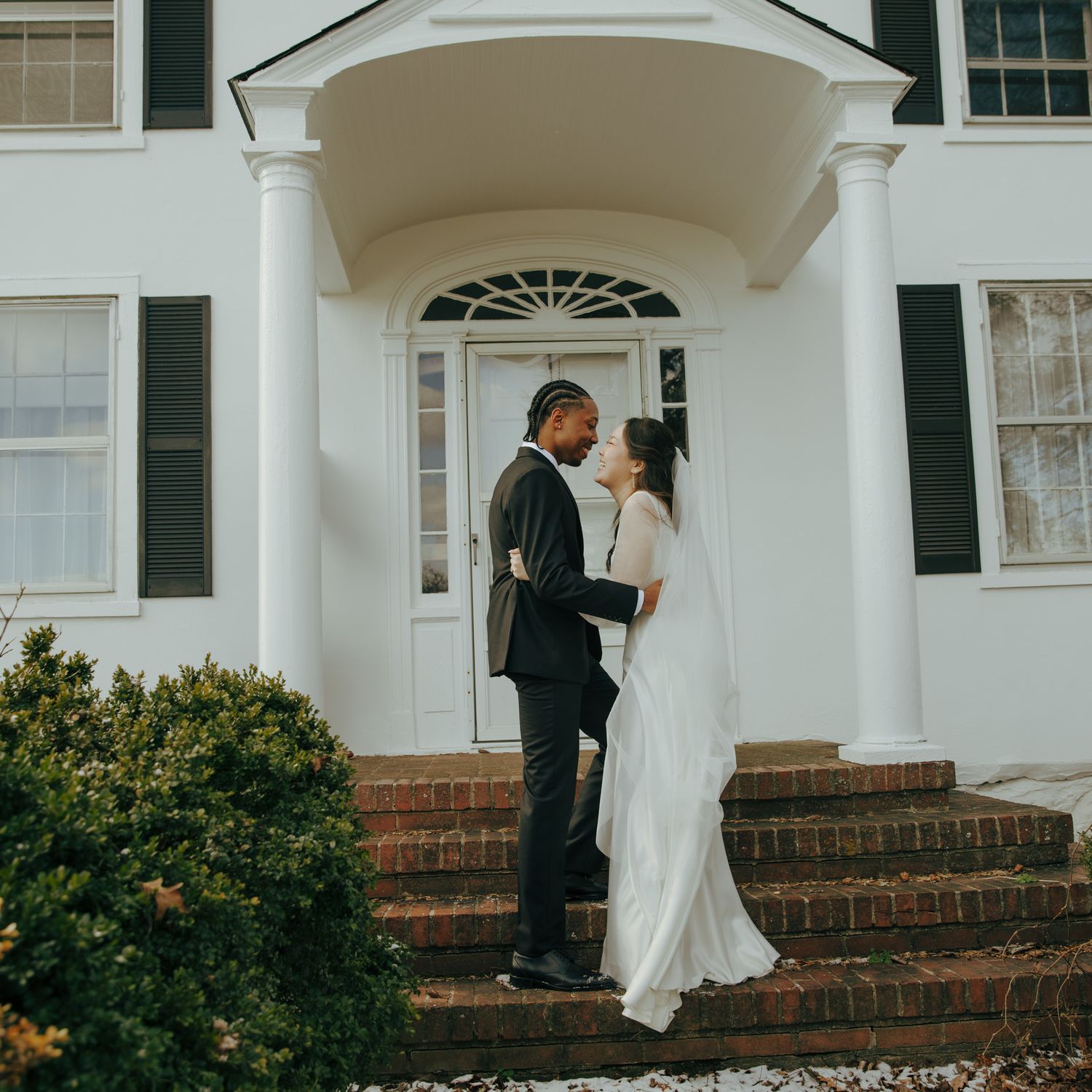 A couple shares an intimate moment on the steps of a grand white colonial house.