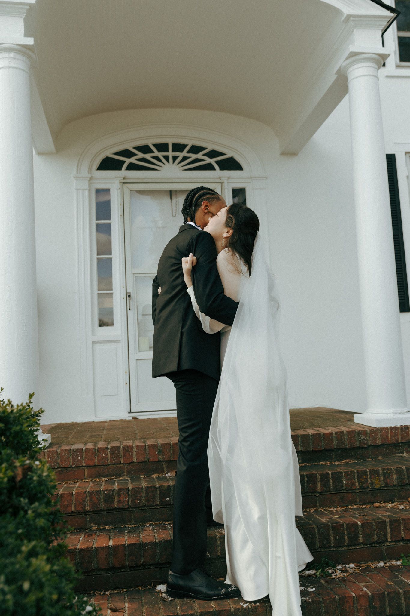 A wedding couple embracing under an elegant white archway entrance with columns.