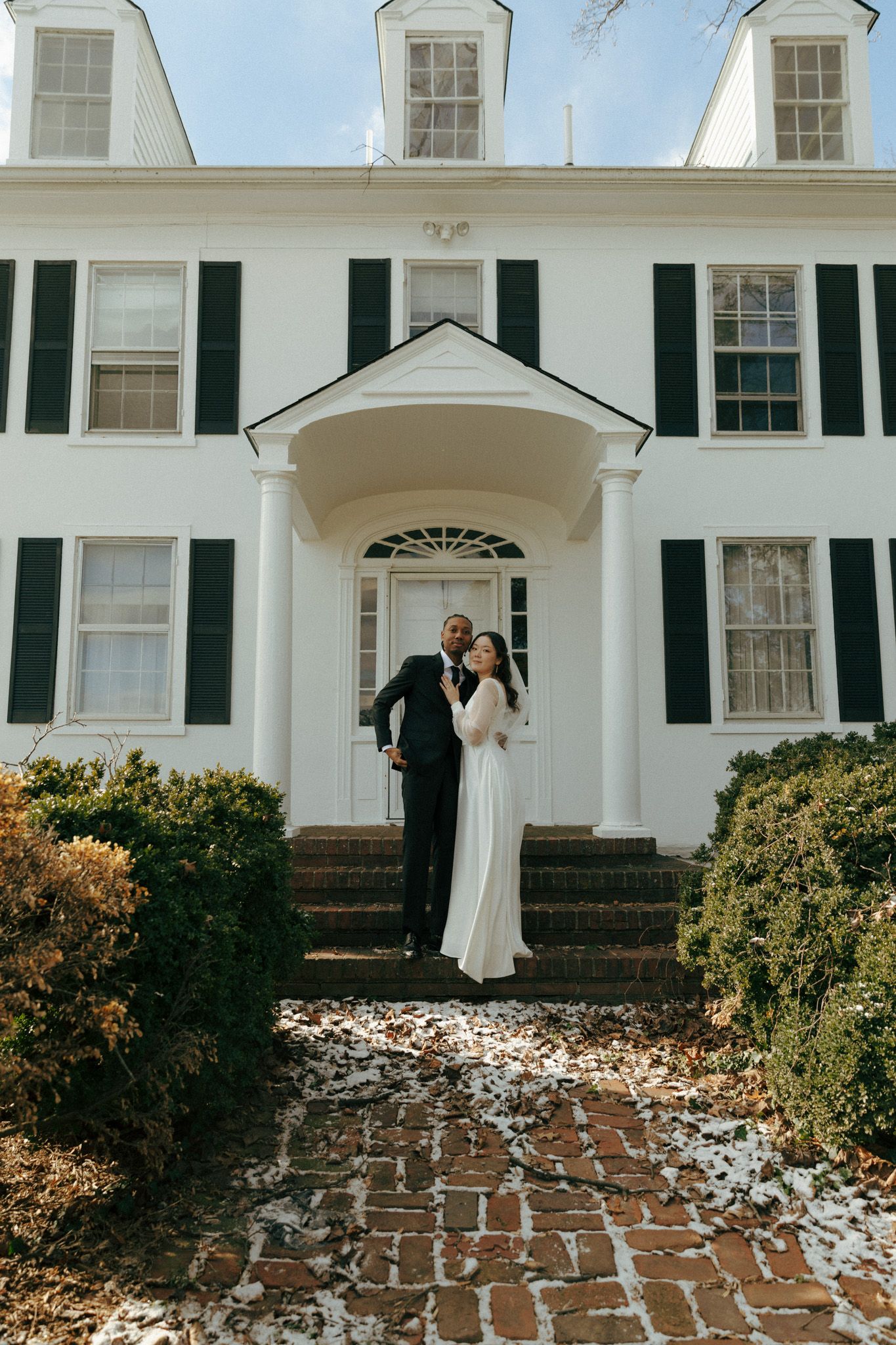 A newlywed couple poses on the steps of a stately white mansion with black shutters.