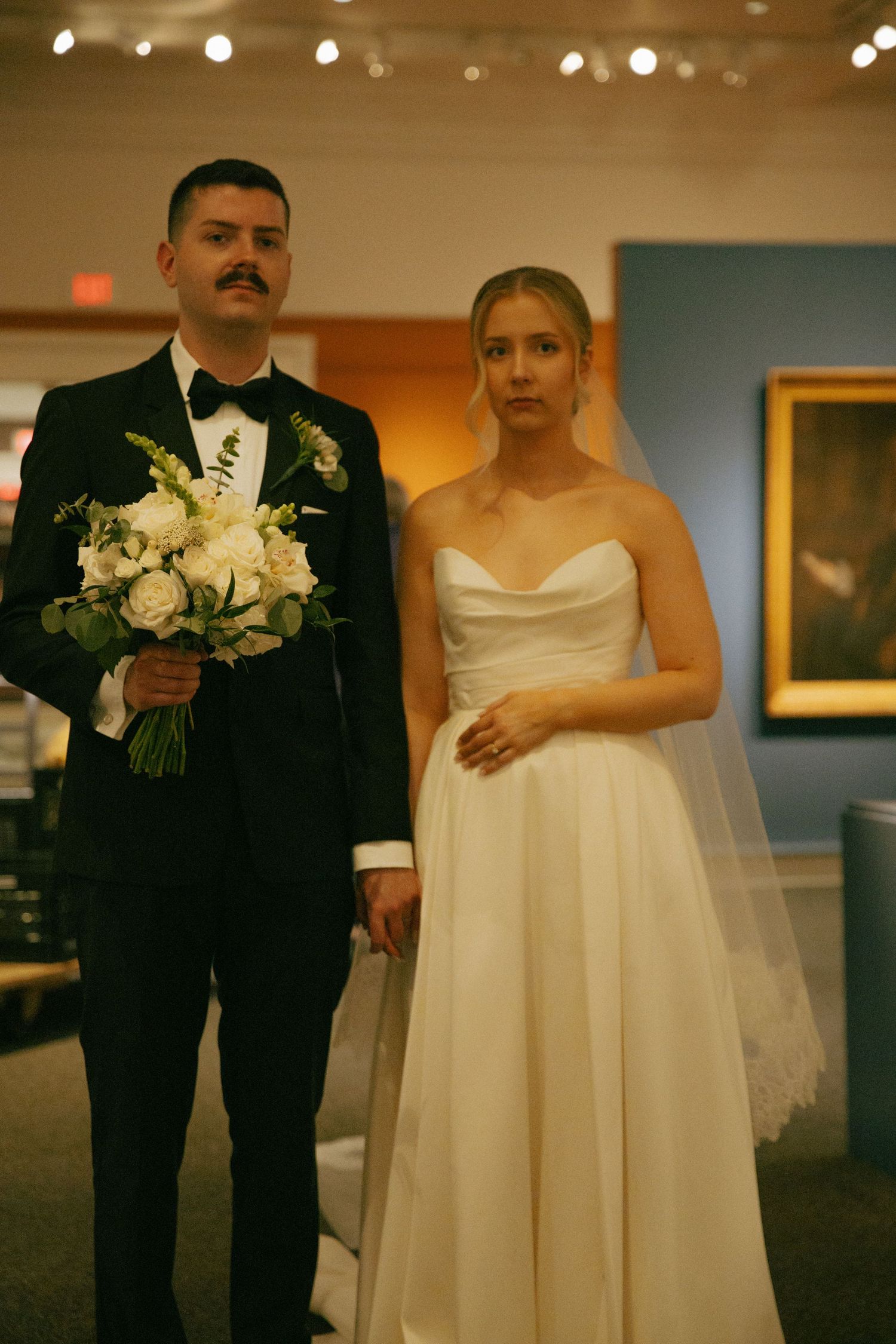 Wedding couple poses formally in art gallery holding white floral bouquet.