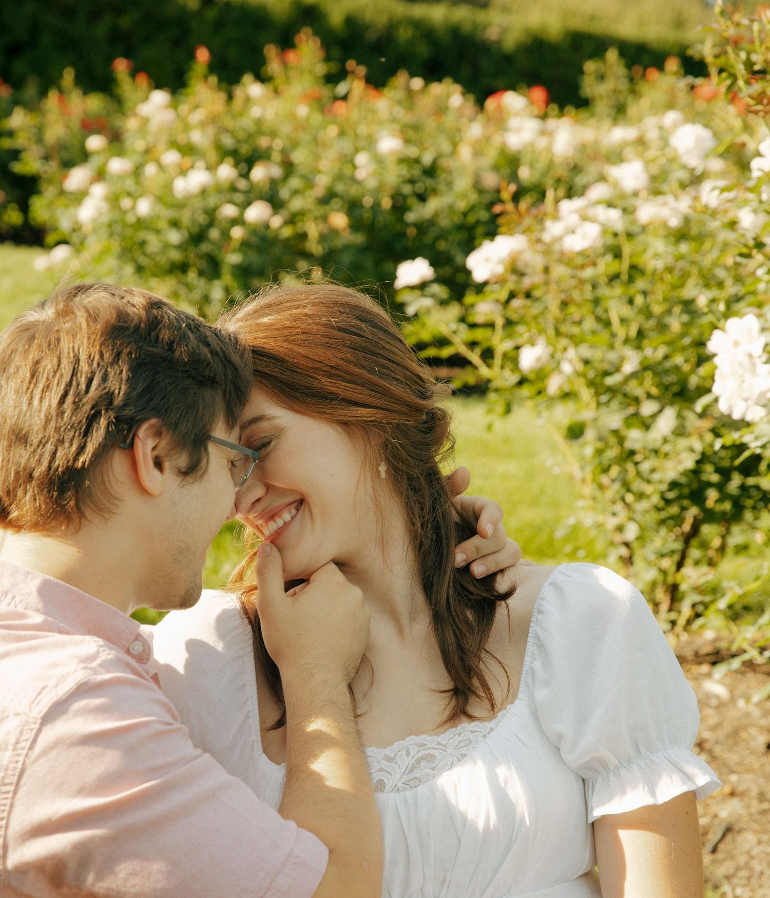 Intimate moment shared between couple in white and pink attire amid flowering garden.