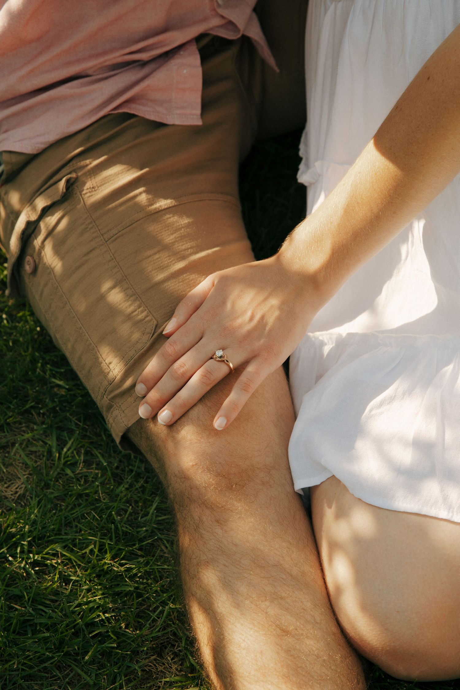Close-up of hands showing engagement ring in dappled sunlight on grassy ground.