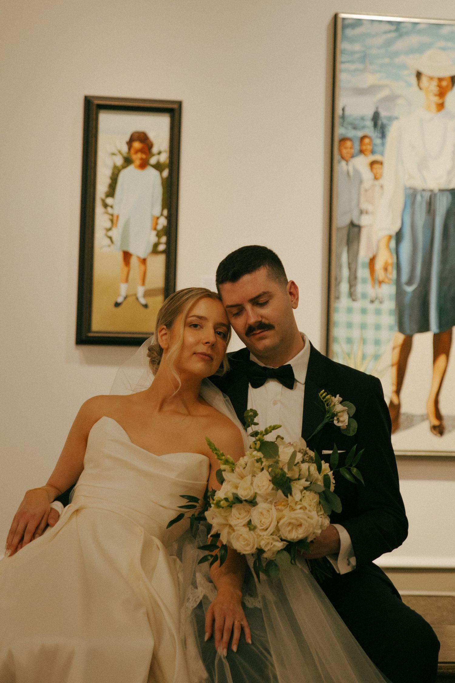 Newlyweds pose casually by vintage portrait paintings in art gallery setting.