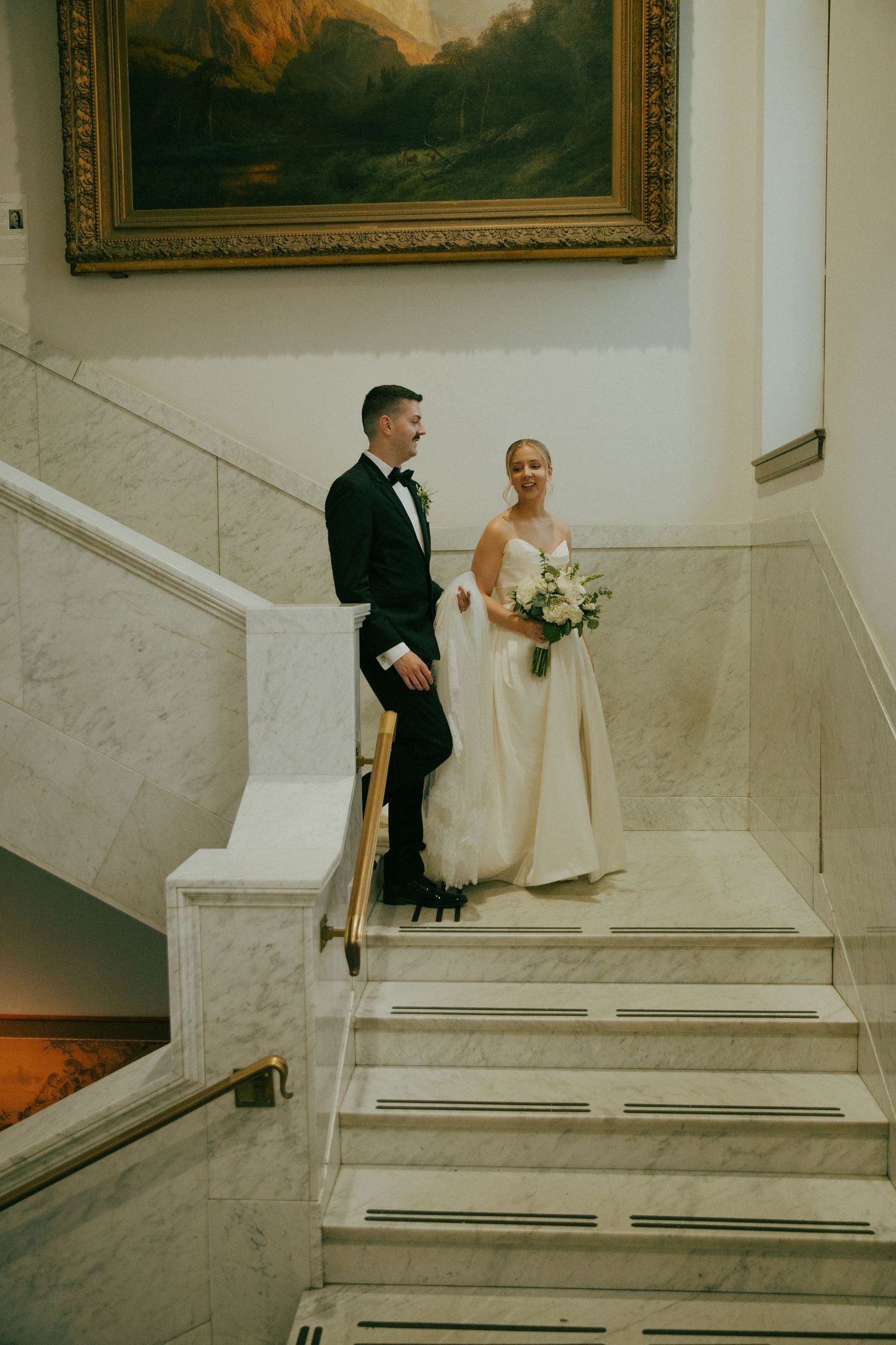 Wedding couple stands elegantly on marble staircase beneath ornate framed artwork in formal attire.