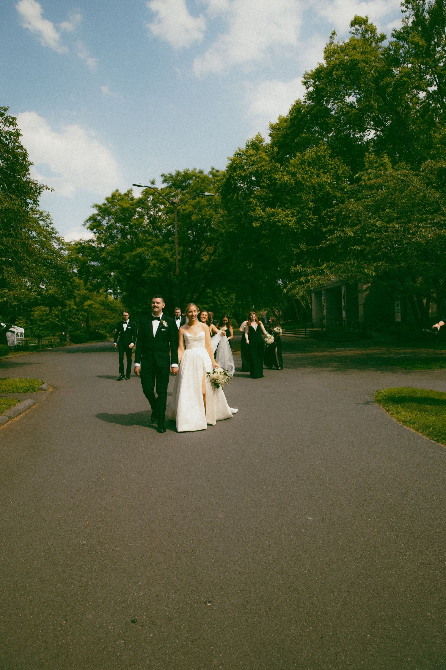 Wedding party walking down outdoor path with trees and blue sky.