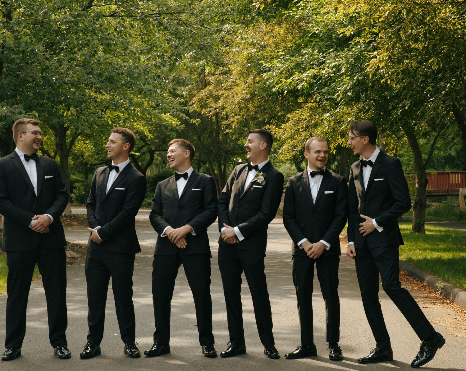 Six groomsmen in matching tuxedos share a candid moment on a tree-lined path.