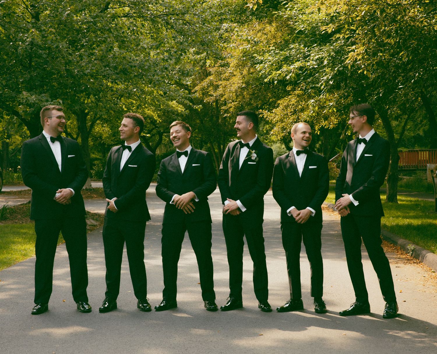 Groomsmen in black tuxedos standing in line on garden path with autumn trees.