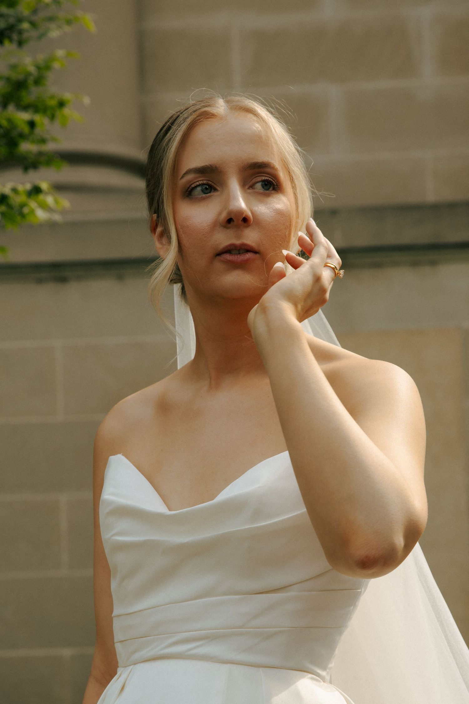 Elegant bride in strapless white gown poses against stone wall with greenery.