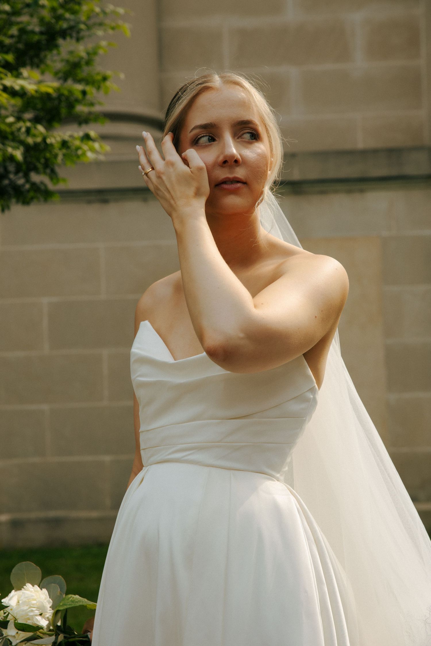 Bride in classic white dress touches face while posing against stone wall.