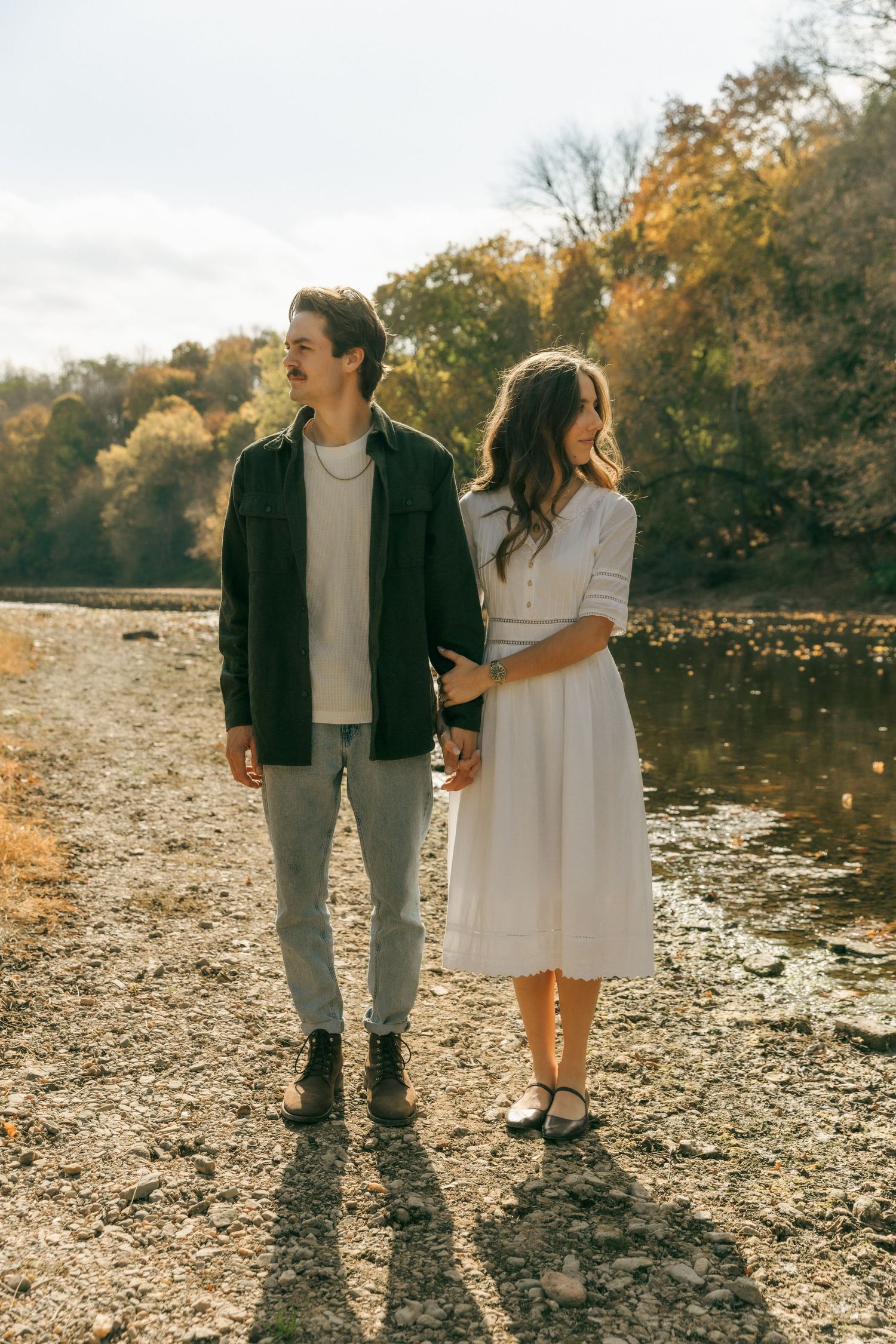Couple standing by a lake during fall with golden trees in background.