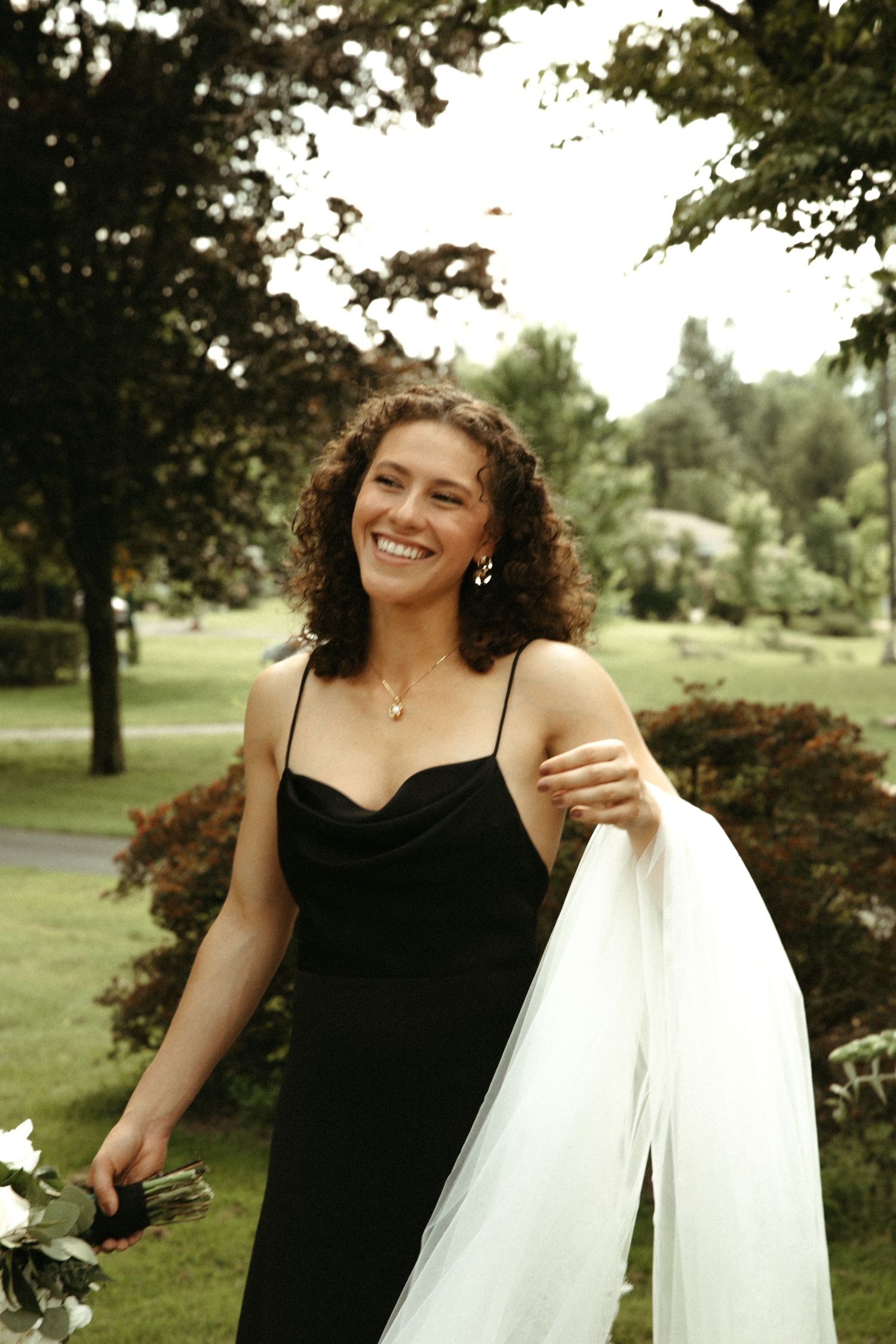 Bridesmaid in black dress holding white veil while smiling in outdoor garden setting.