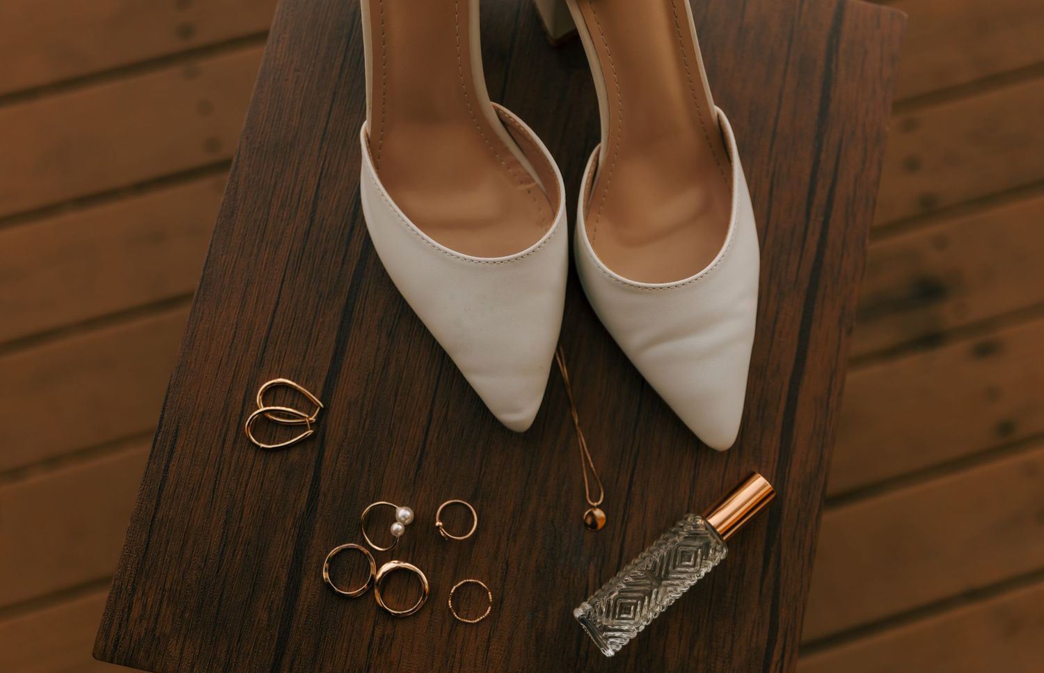 White pointed-toe wedding shoes with rings and accessories on dark wooden surface.