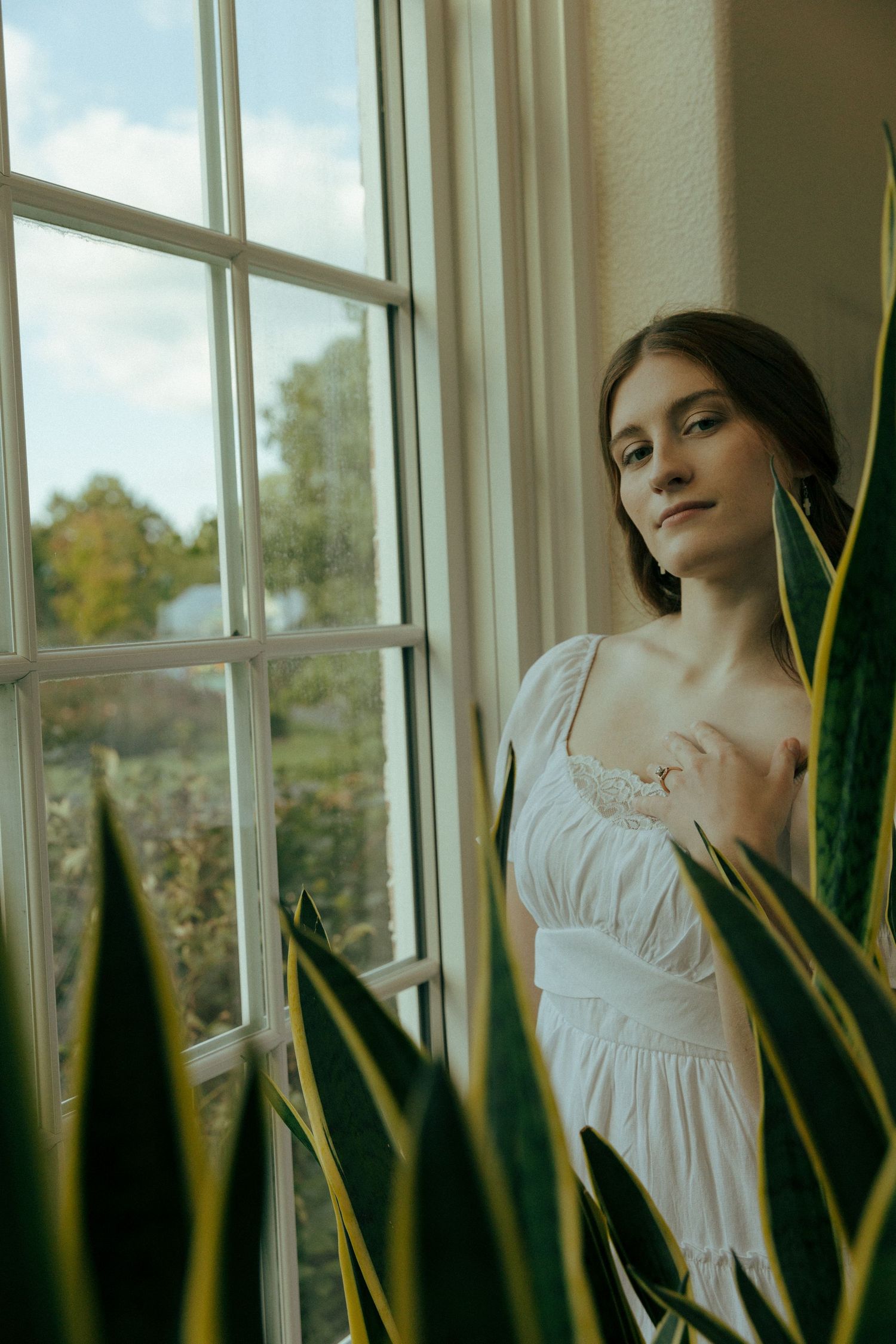 Person in white dress gazes through window framed by indoor plants.