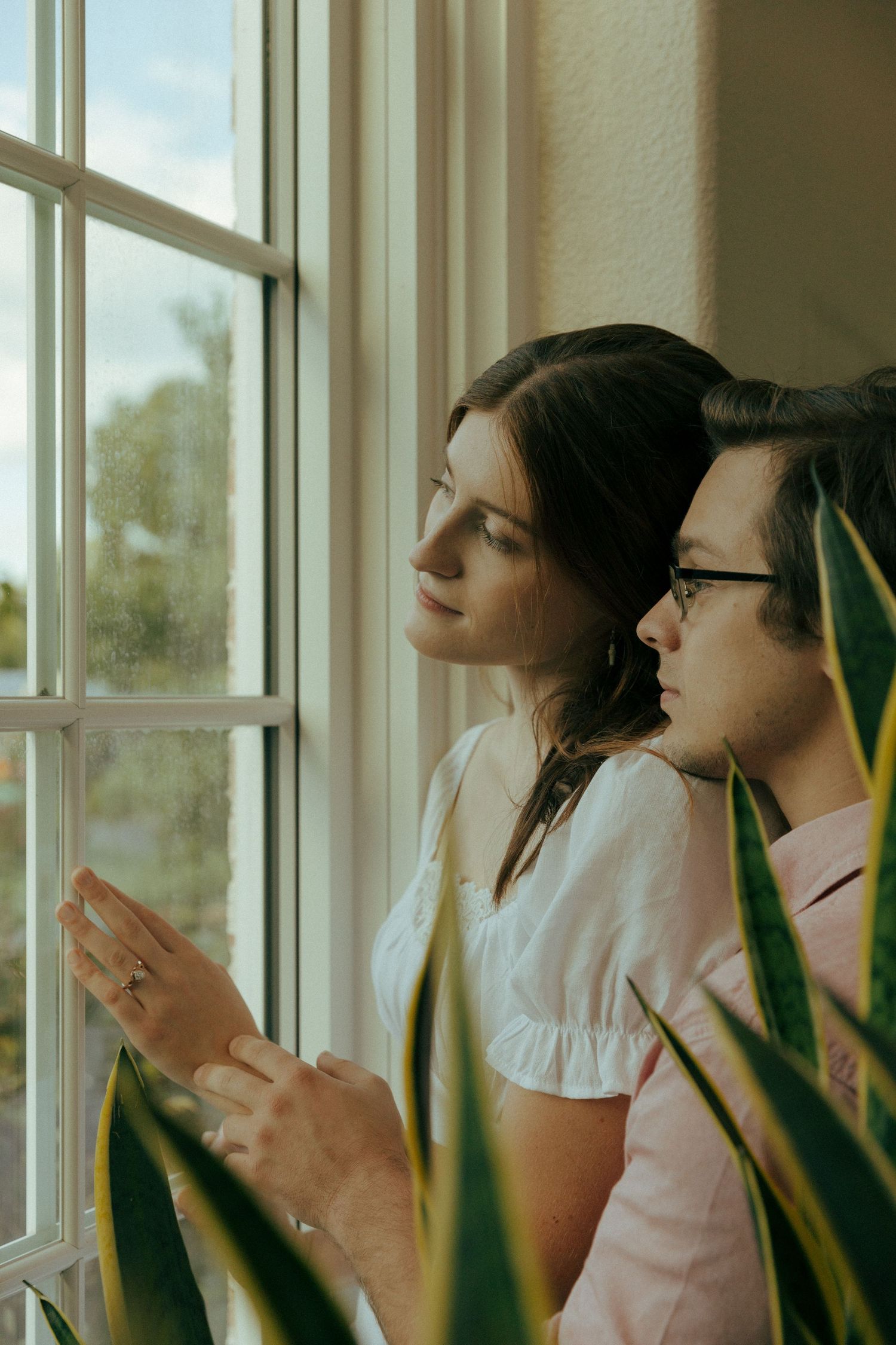 Two people gaze out a sunlit window with house plants in foreground.