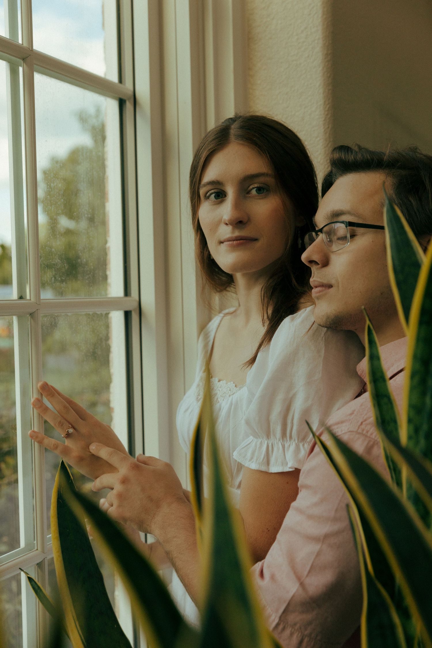 Couple standing by window with snake plants in romantic indoor photoshoot.