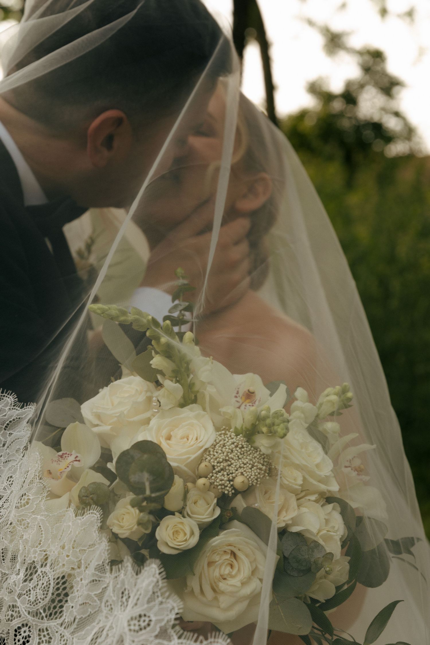 White rose wedding bouquet held between couple under delicate lace veil.