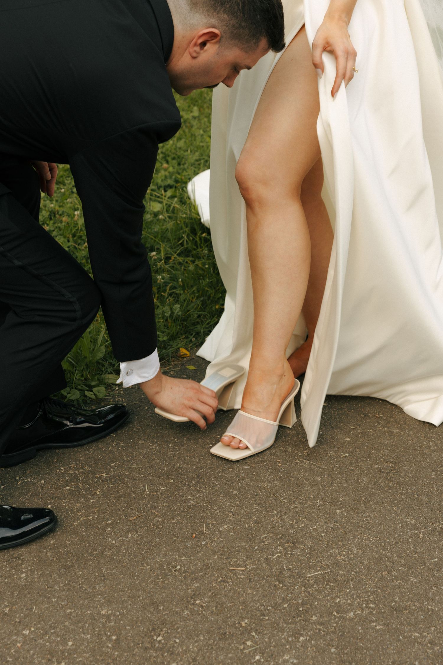 Close-up of someone helping adjust a bride's white high-heeled shoe on a path.