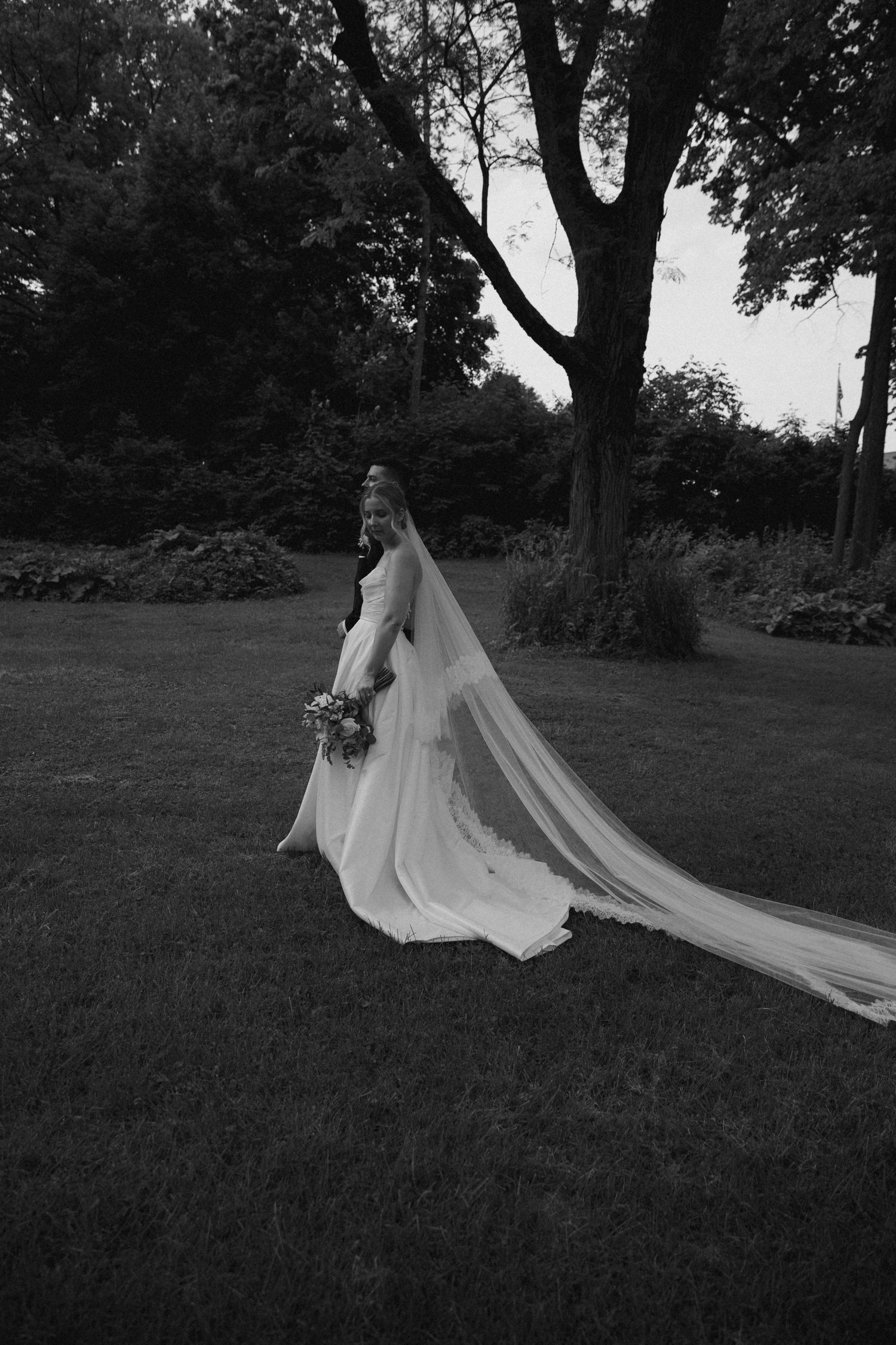 A black and white photo of a bride with flowing veil standing in a garden setting.