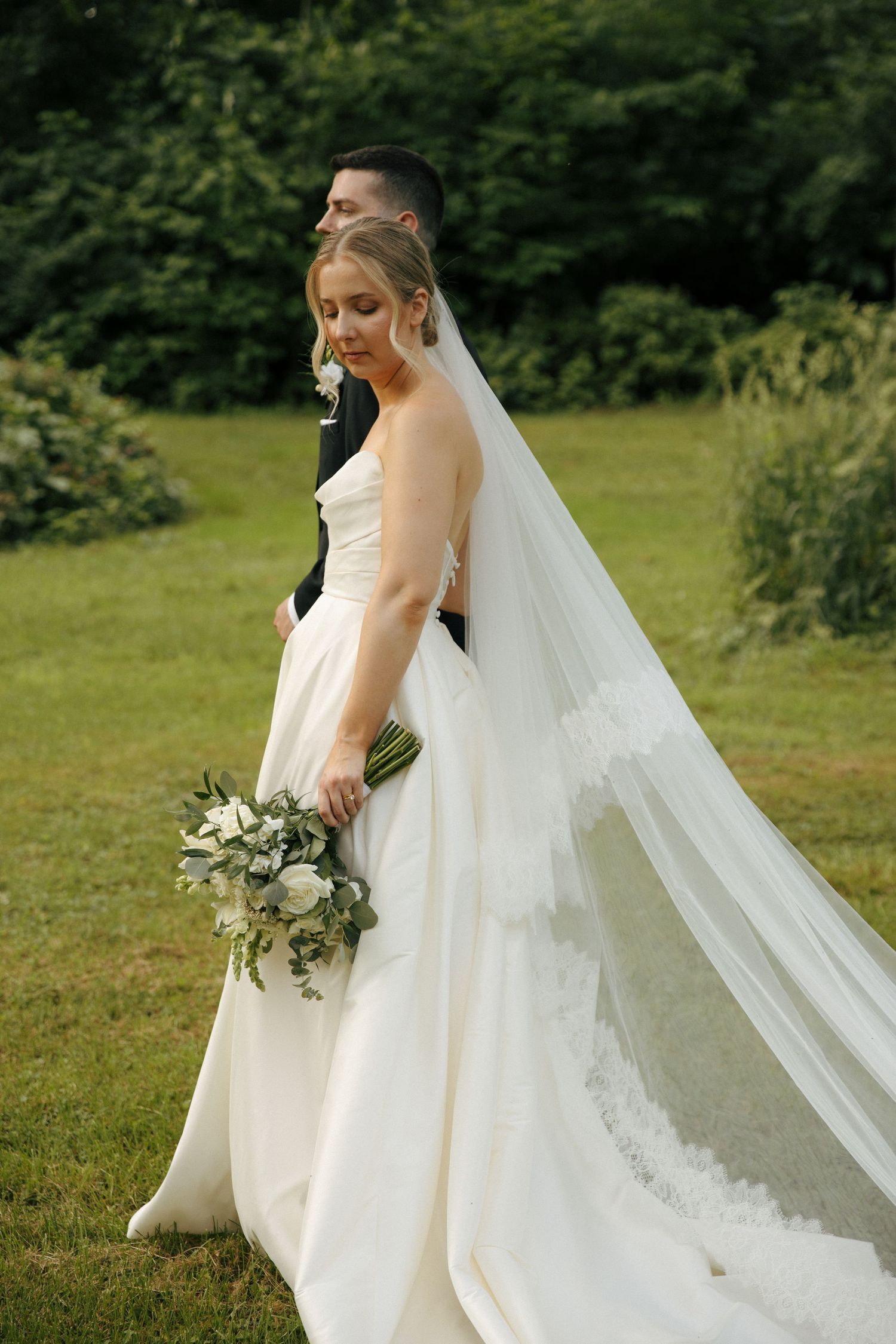 A bride in an elegant white gown holds a bouquet while standing on grass with trees in background.