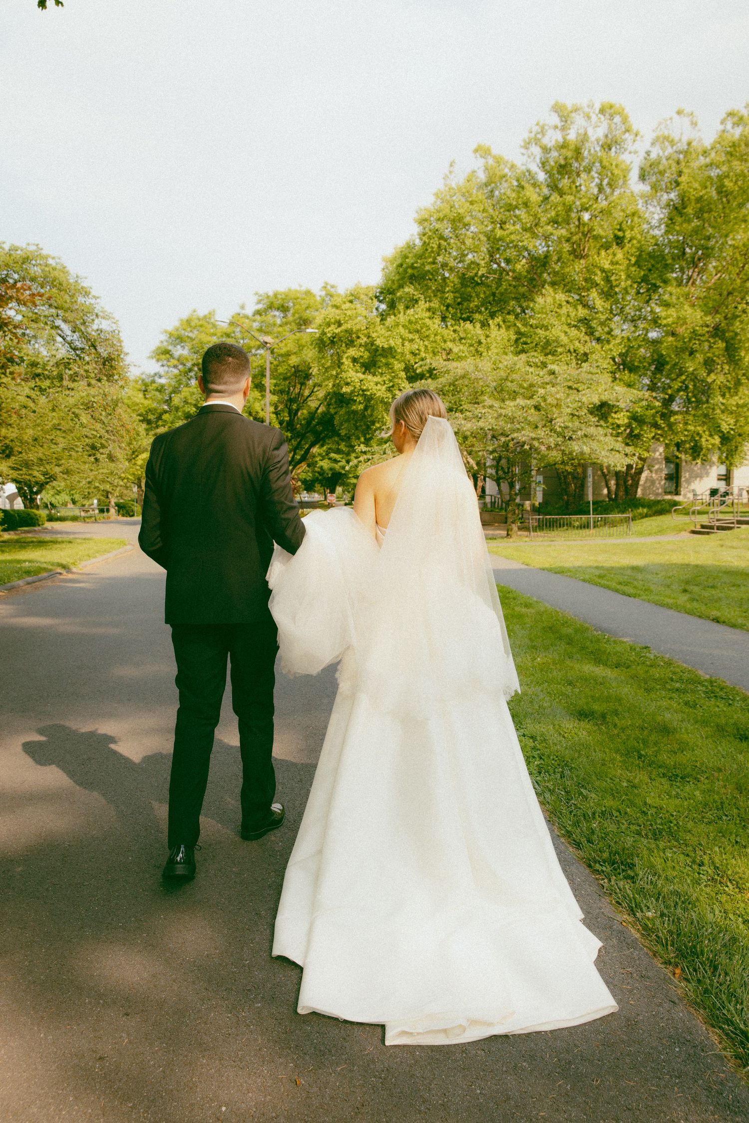 A wedding couple walks along a tree-lined path, with the bride's white dress and veil trailing behind.