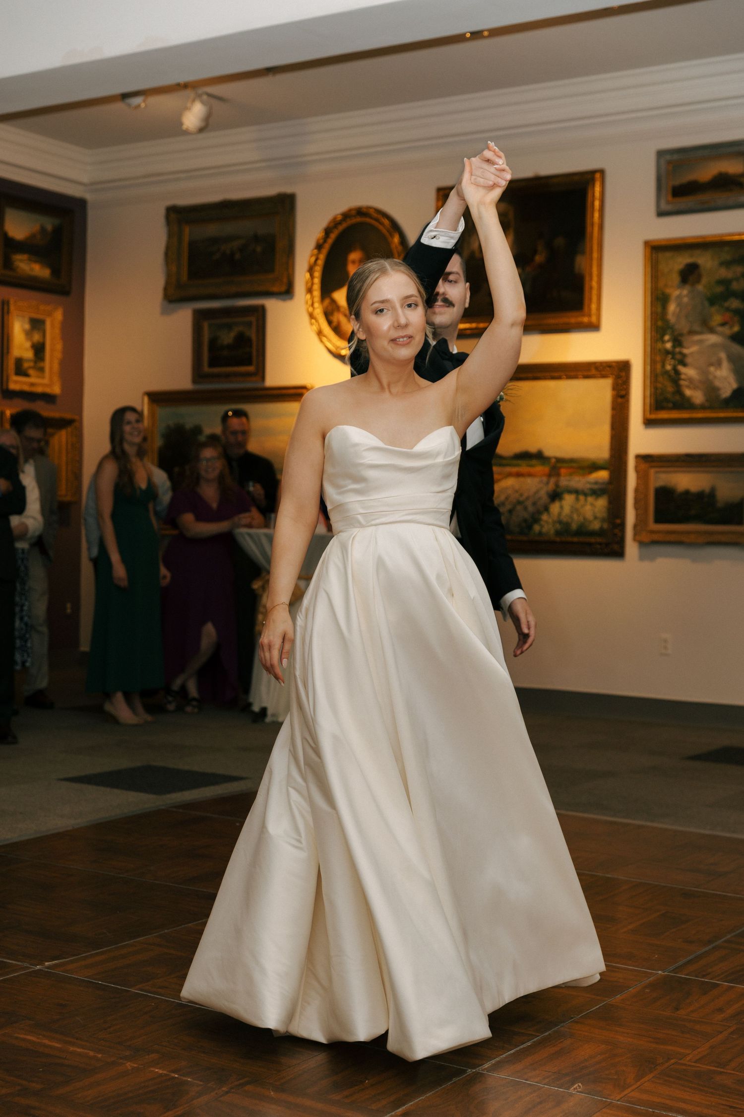 A bride in a flowing white gown dances gracefully in an art gallery setting.