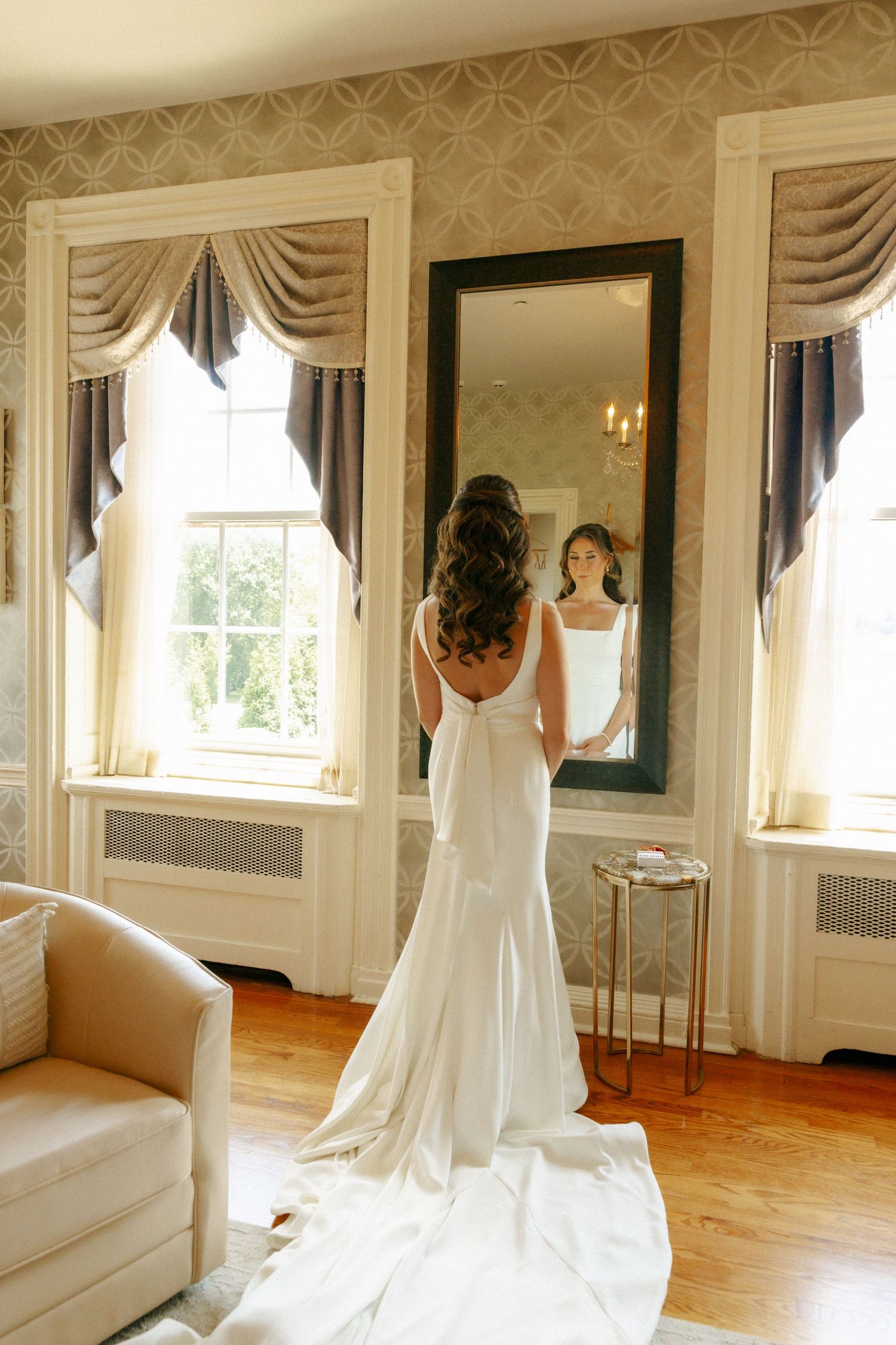 Bride in elegant white gown views her reflection in a large mirror in a beautifully decorated bridal suite.
