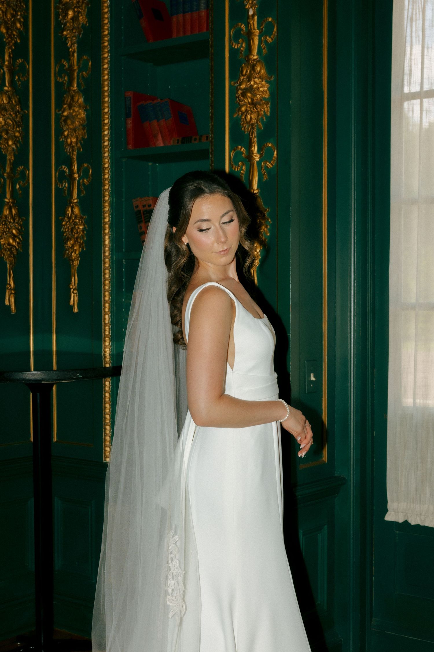 Bride in elegant white satin gown and lace veil poses in a room with green walls and gold ornate details.