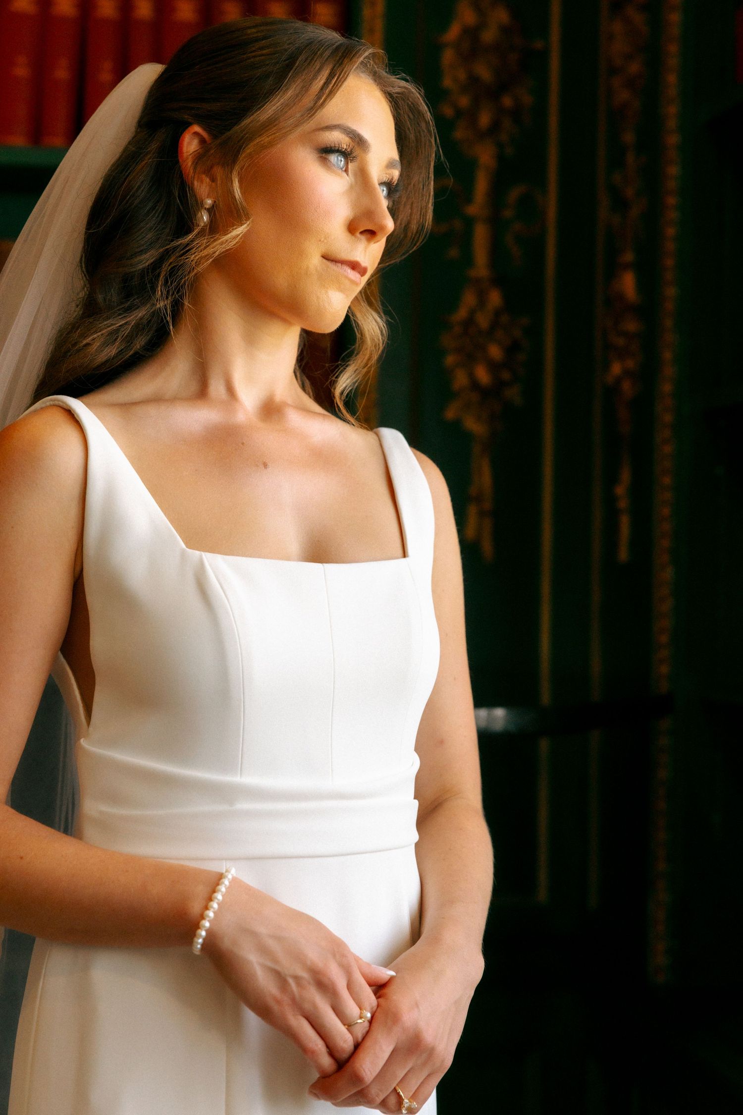 Elegant bride in a white square-neck gown with veil, pearl bracelet, and soft makeup, posing in a dark ornate room.