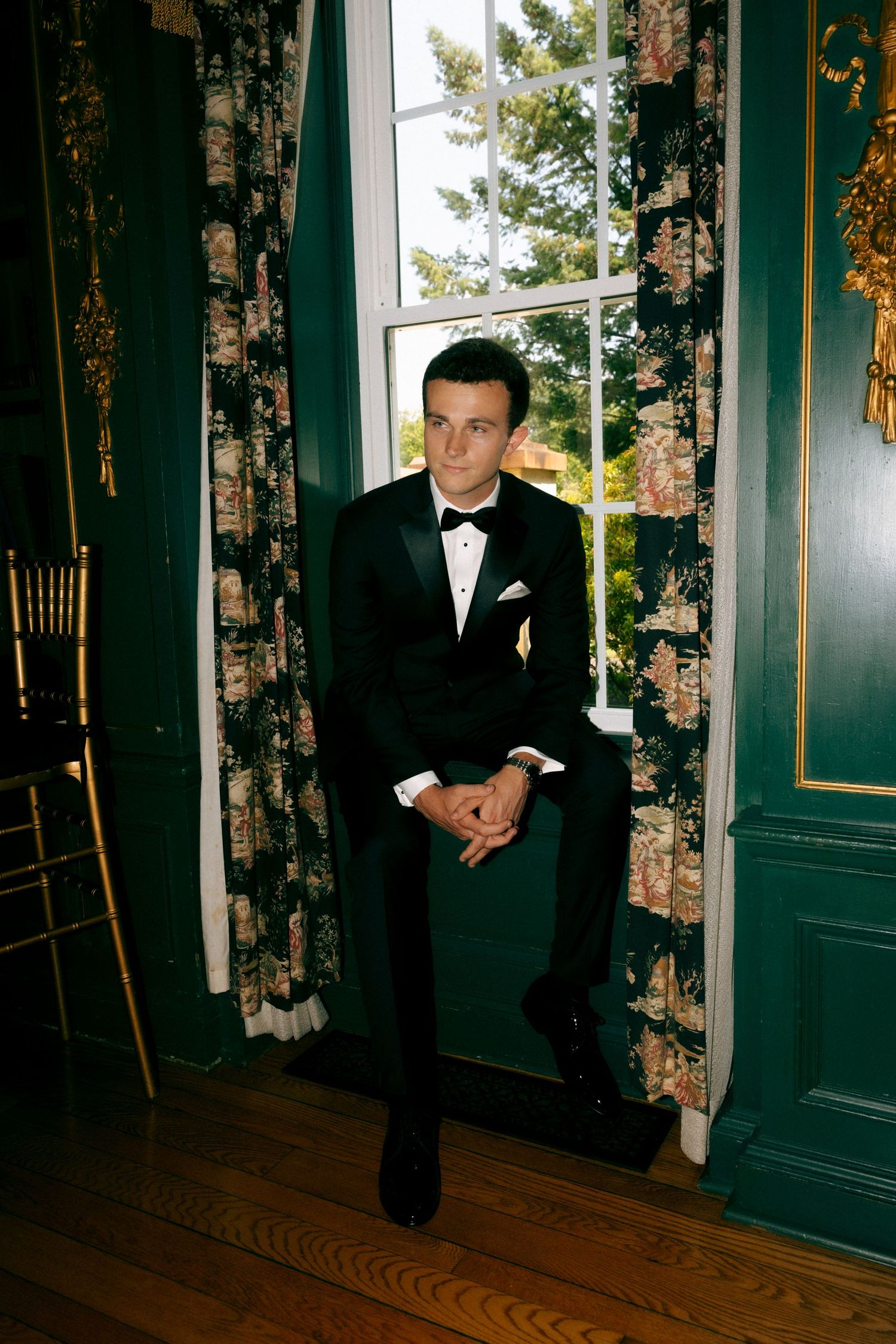 A man in a black tuxedo with bow tie sits by a window with floral curtains in an elegant green room.