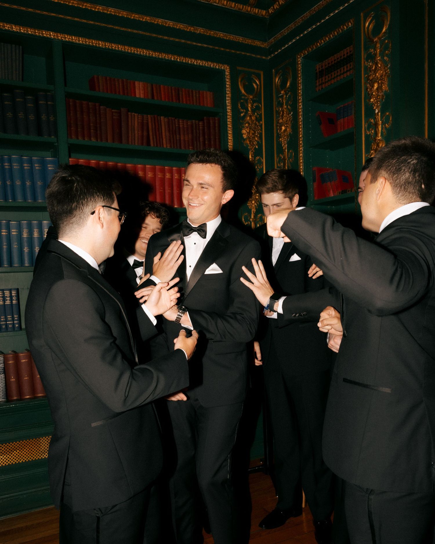 Men in black tuxedos socializing in an elegant library room with green shelves and gold trim.