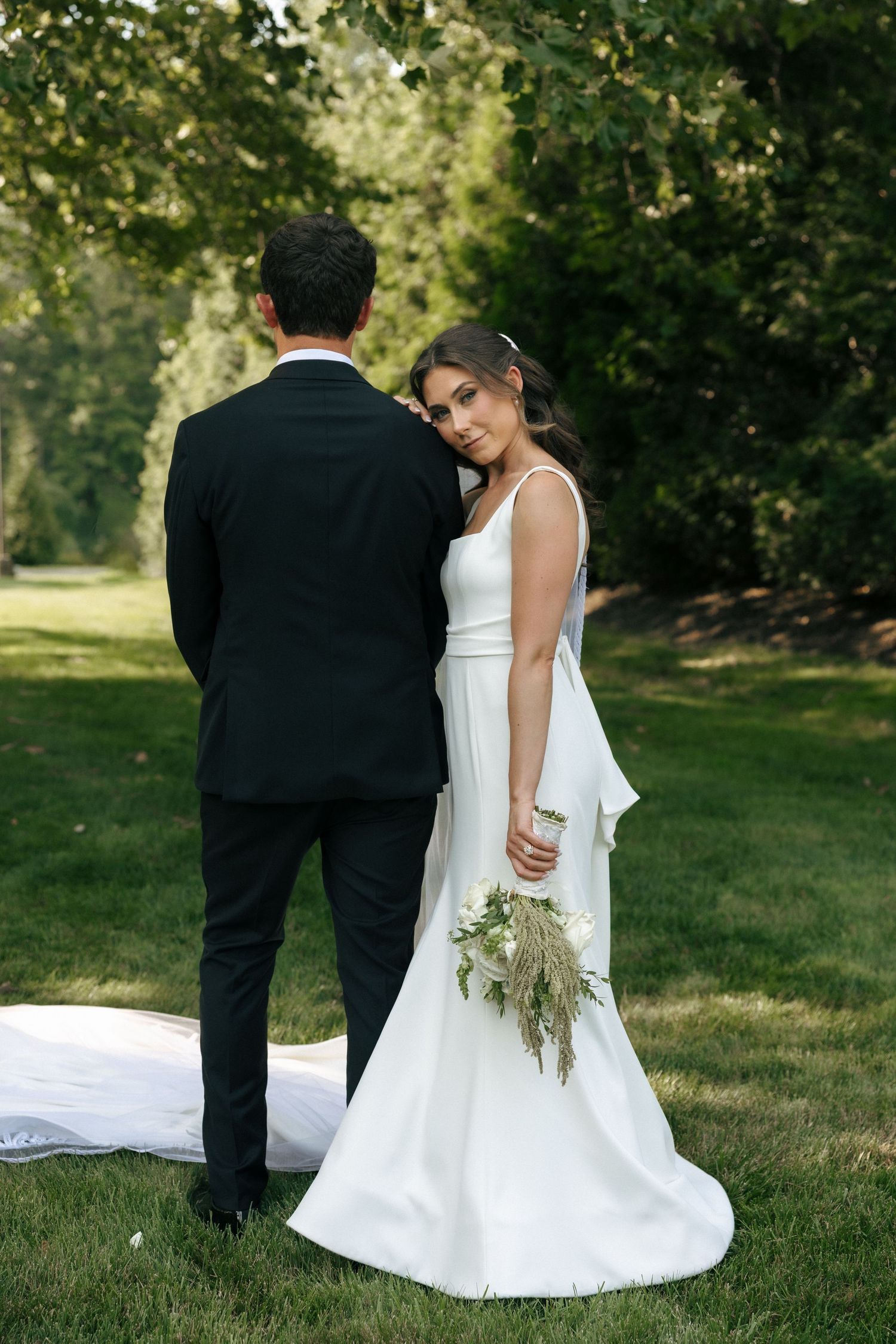 Bride in white gown holds bouquet beside groom in black suit, posing outdoors on lush green lawn.