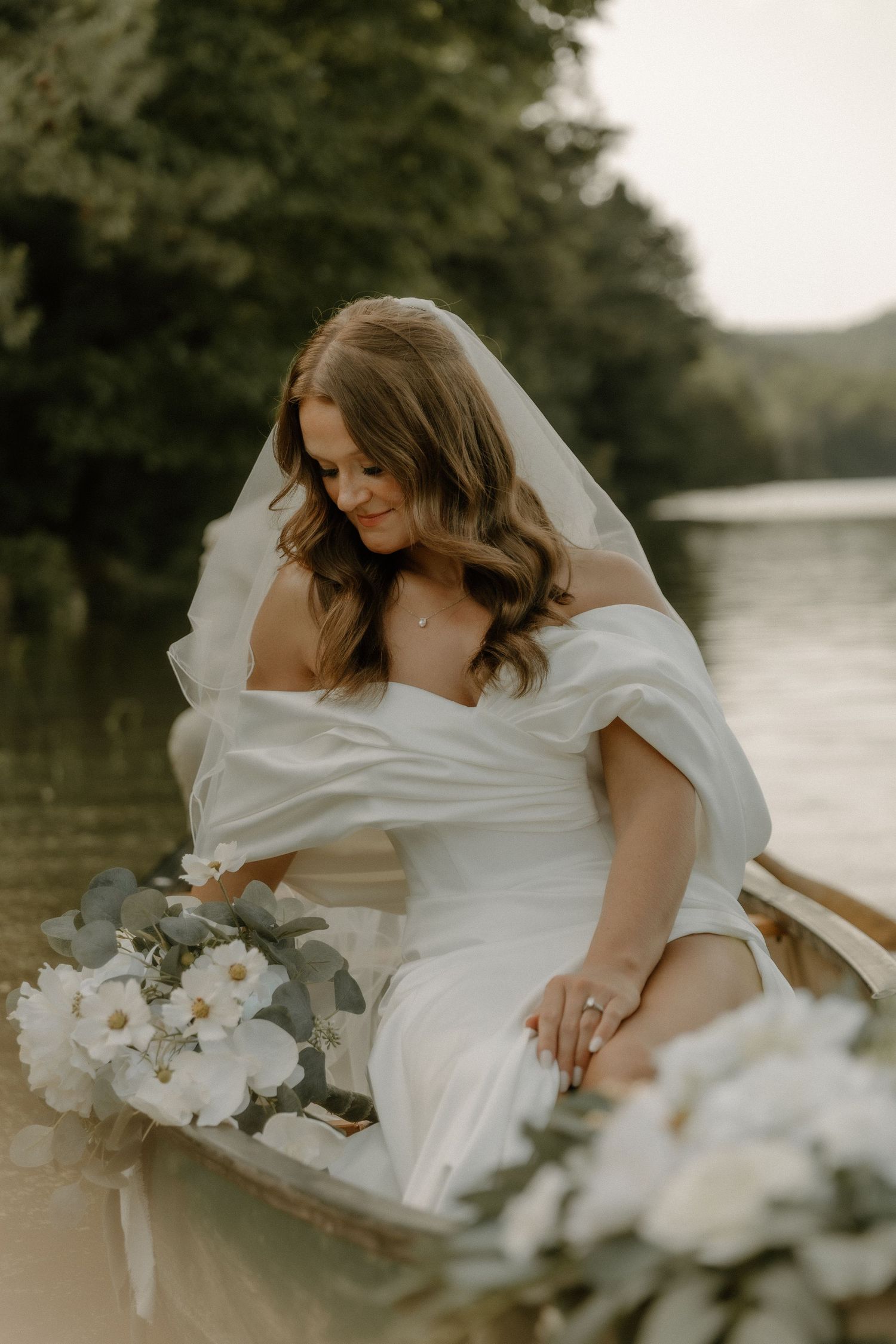 A bride in a white dress sits in a canoe on a lake, holding a bouquet of white flowers.