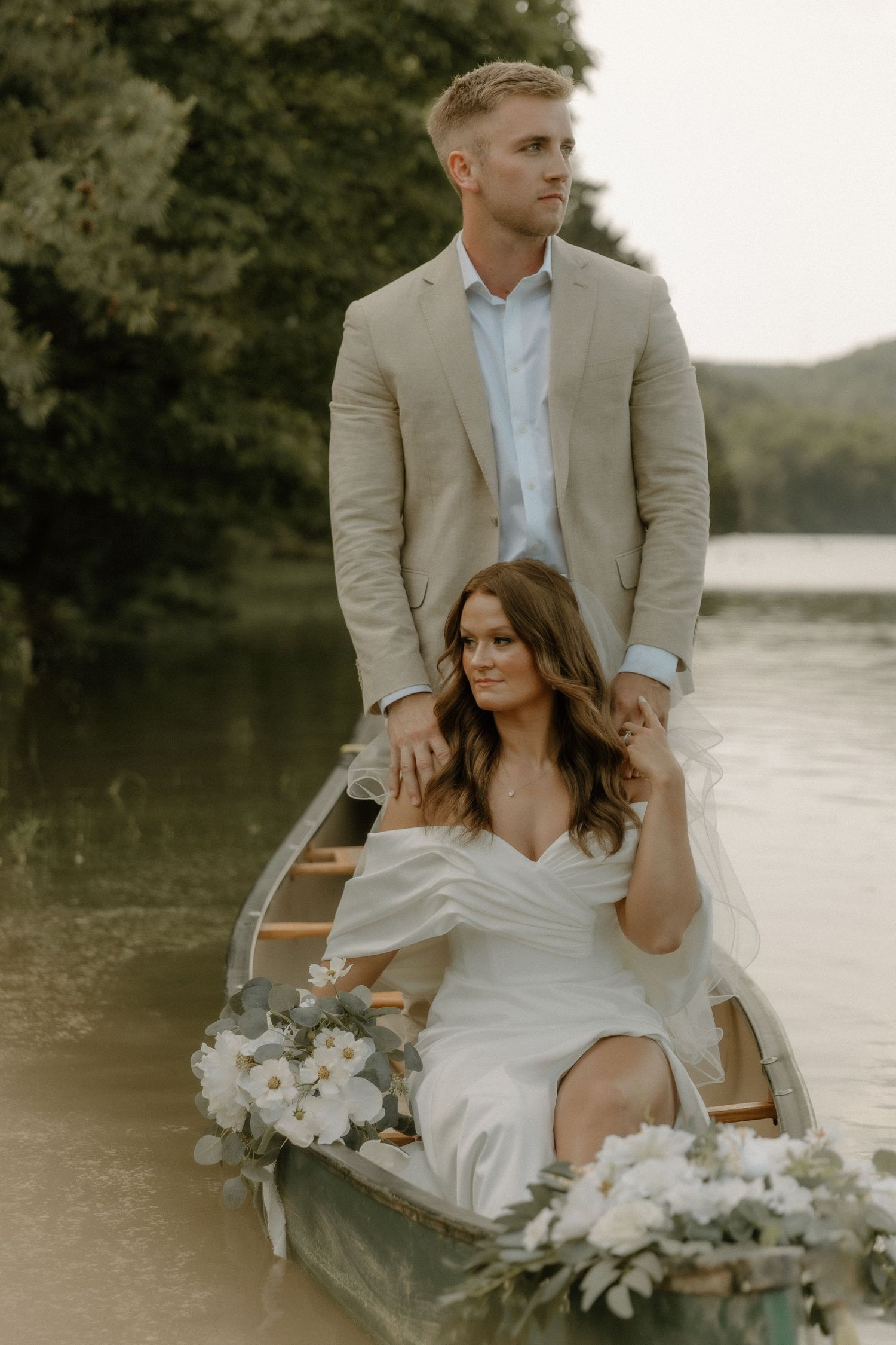 Elegant couple posing in a canoe decorated with white flowers on a peaceful lake.