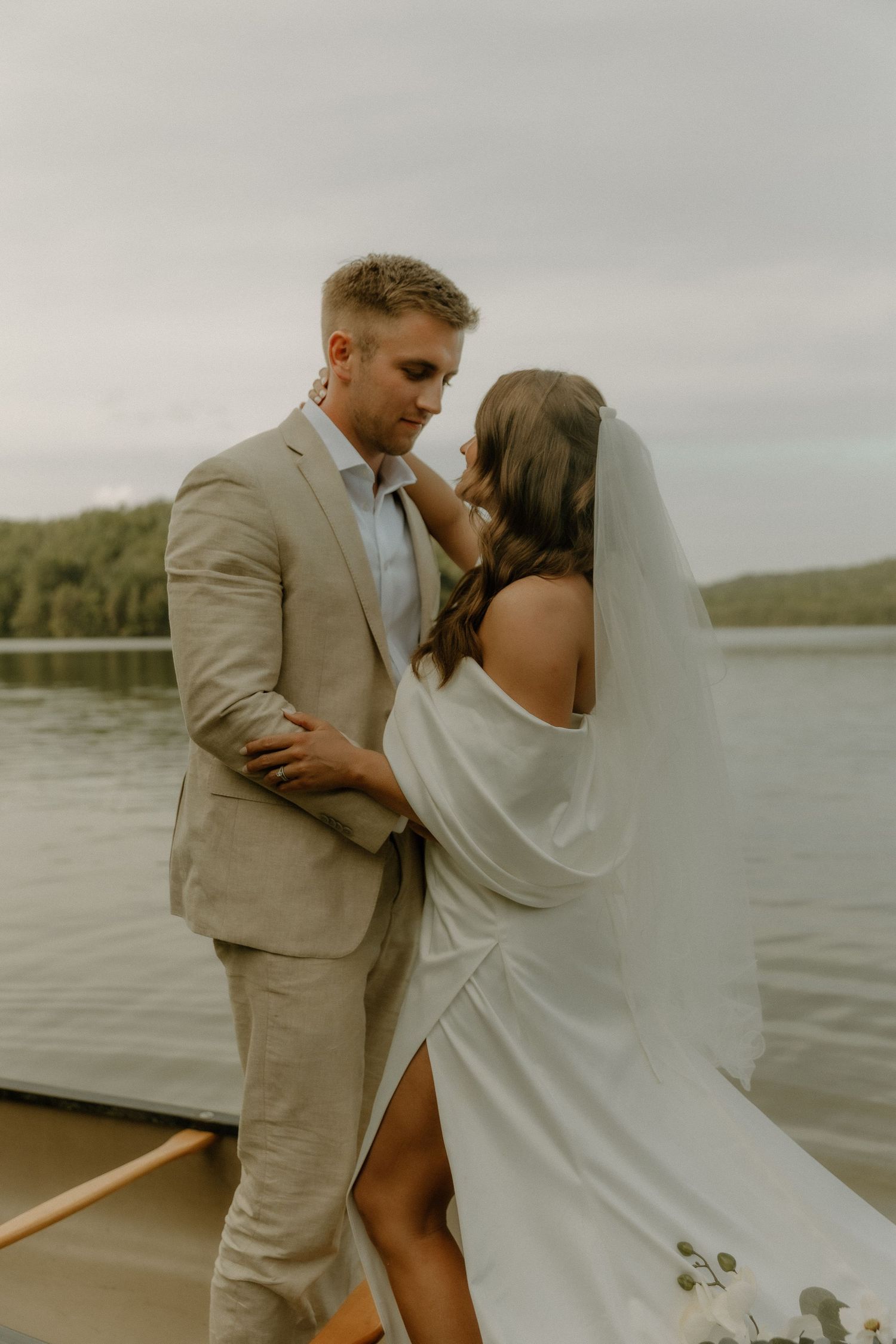 A romantic wedding couple embraces in a boat on a serene lake surrounded by trees.